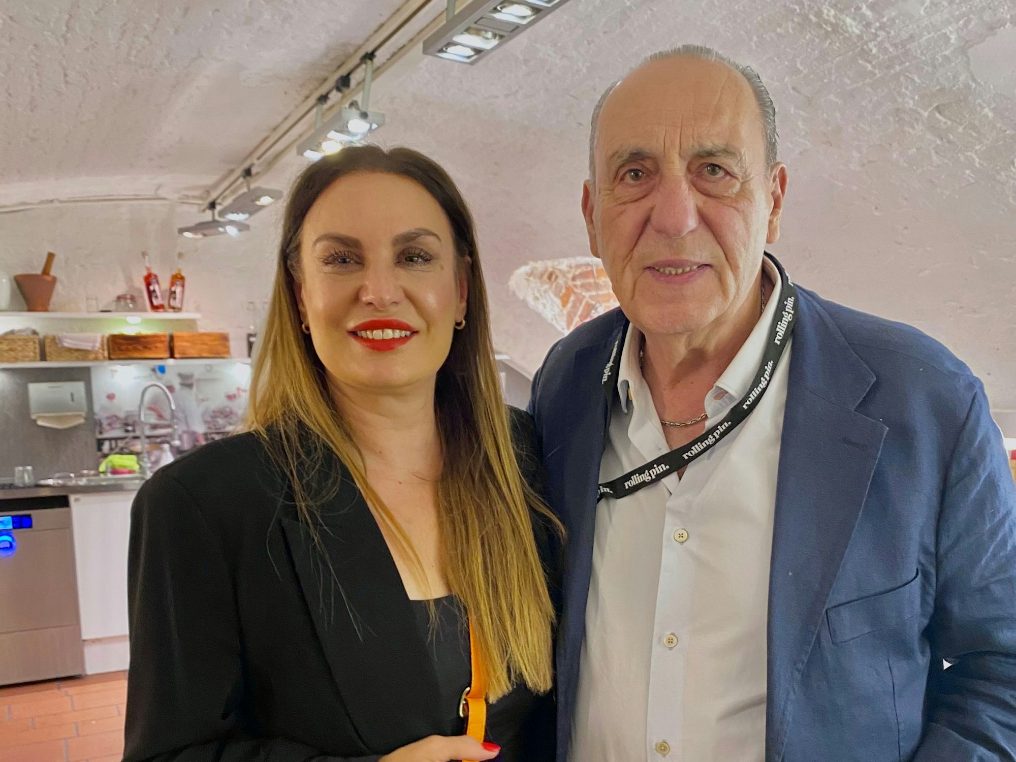 Eine Frau mit langem Haar und schwarzer Kleidung und ein älterer Herr im Anzug mit weißem Hemd stehen in einer Küche.