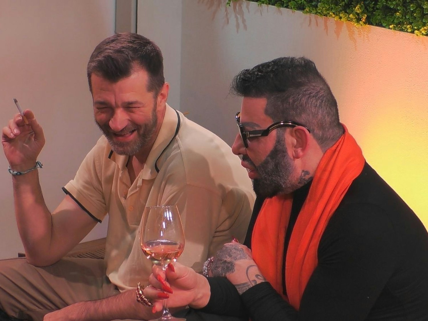 Zwei Männer sitzen nebeneinander, der eine hält ein Glas Wein in der Hand, der andere eine Zigarette.