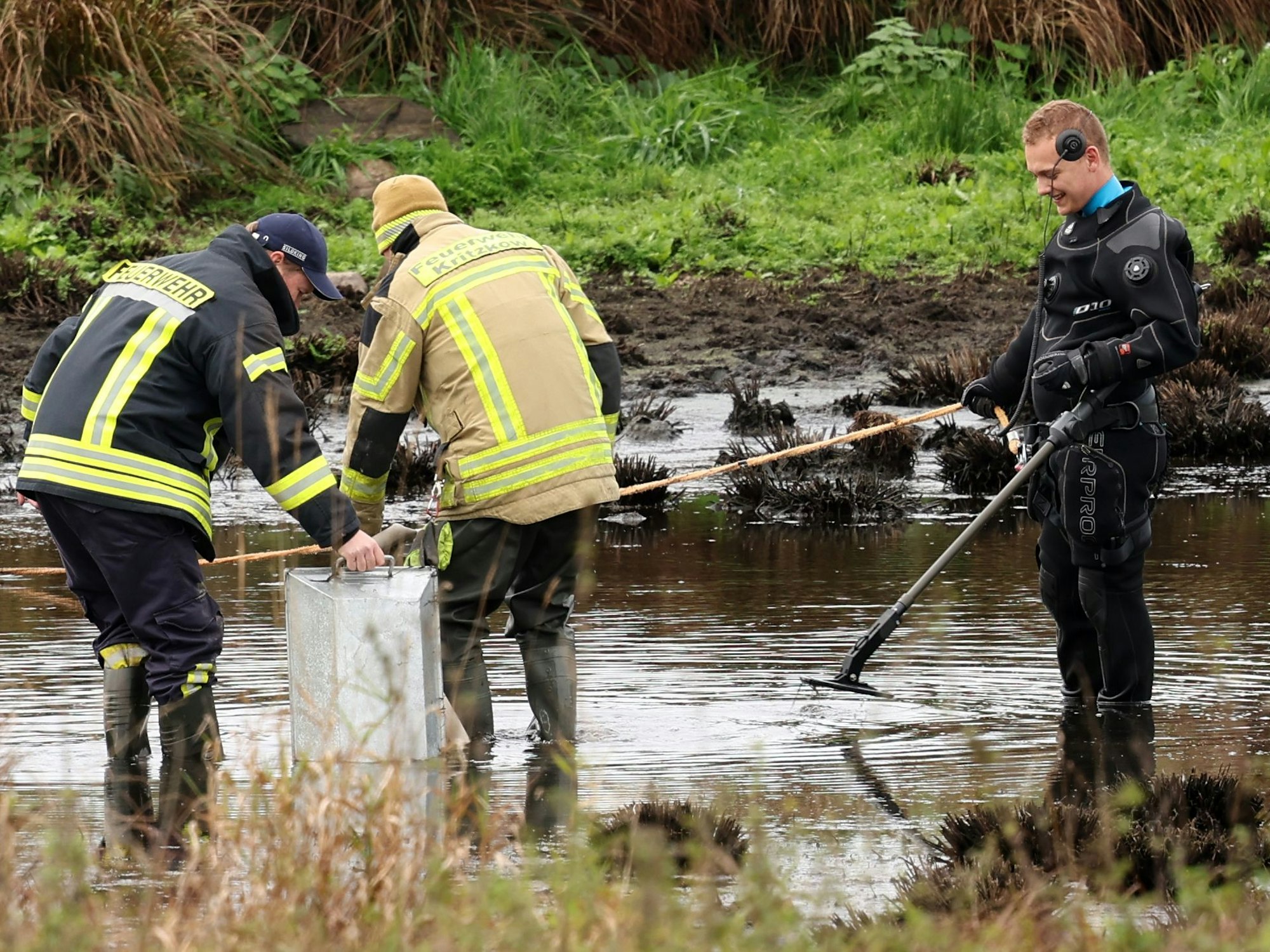 Drei Männer stehen an einem Tümpel, darunter ein Polizist mit einem Detektor.