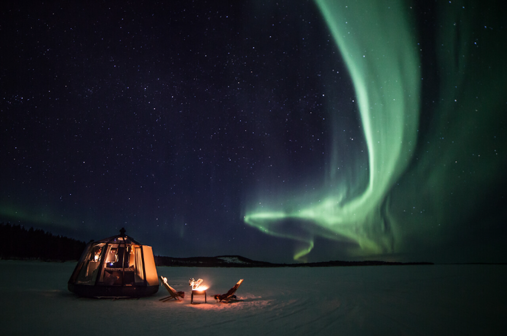 Vor dem dunklen, sternenklaren Himmel leuchten grüne Polarlichter in spektakulären Bögen über eine weite, verschneite Landschaft