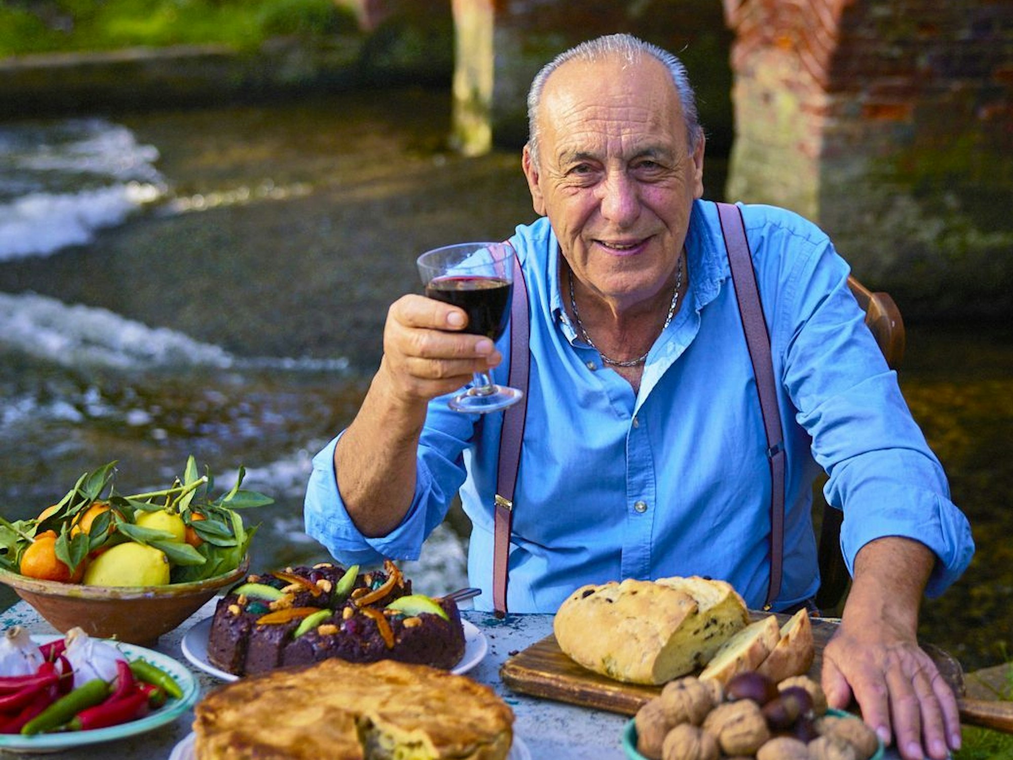 Ein älterer Mann in blauem Oberhemd sitzt vor einem mit Brot, Früchten und Gemüse reich gedeckten Tisch und prostet mit einem Glas Rotwein in die Kamera.