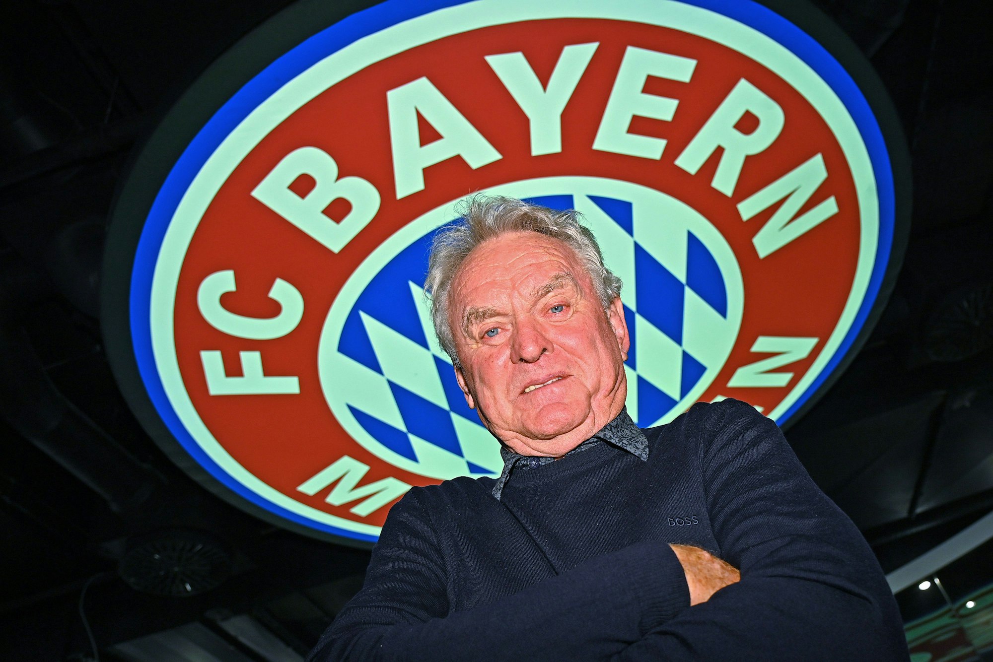 Sepp Maier steht mit verschränkten Armen vor einem Logo des FC Bayern.