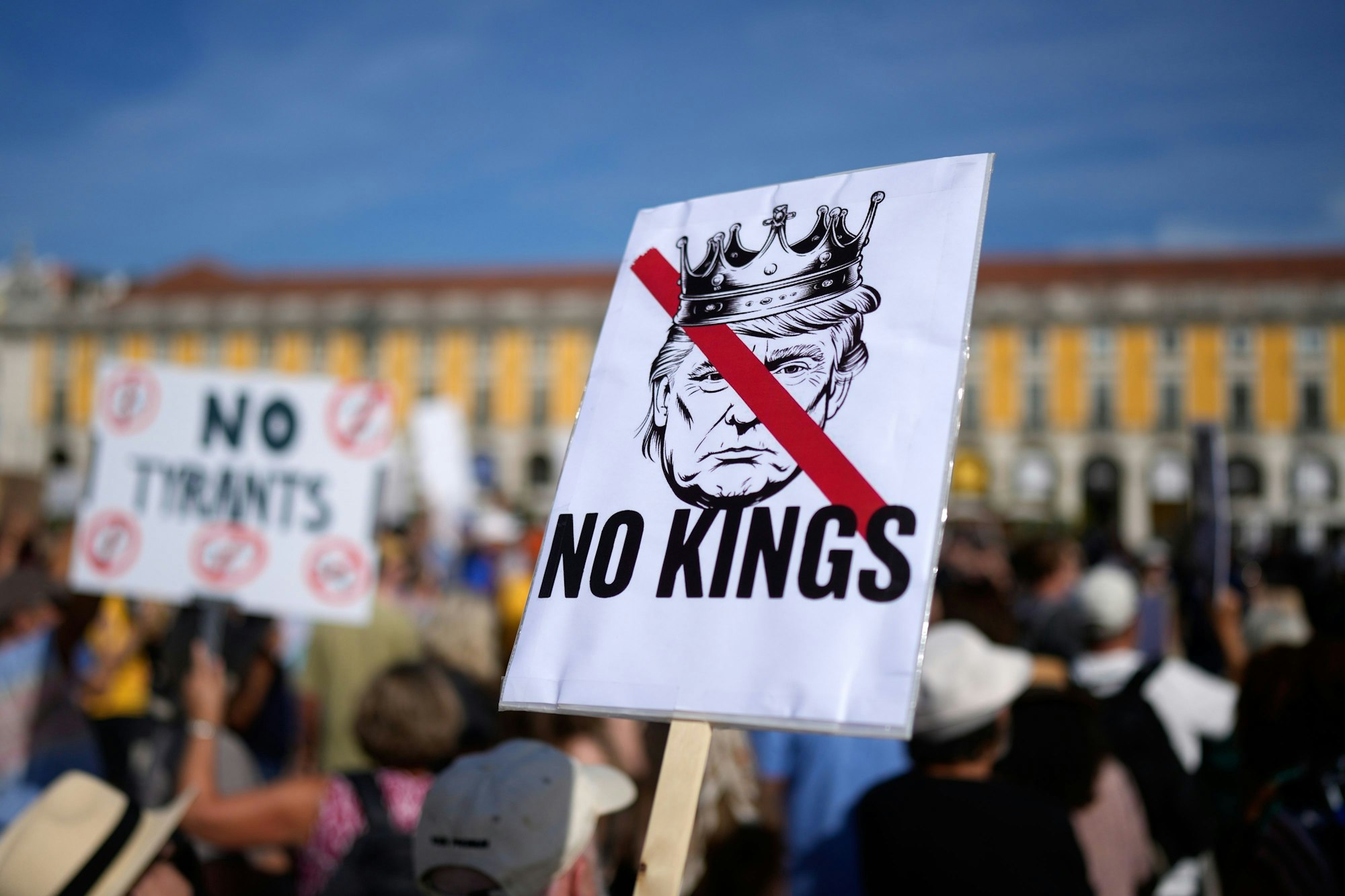 Bei einer Demo hält ein Teilnehmer ein Plakat hoch, das ein gemaltes Porträt von Donald Trump mit einer Krone auf dem Kopf zeigt. Sein Kopf ist durchgestrichen, darunter steht „No Kings“.