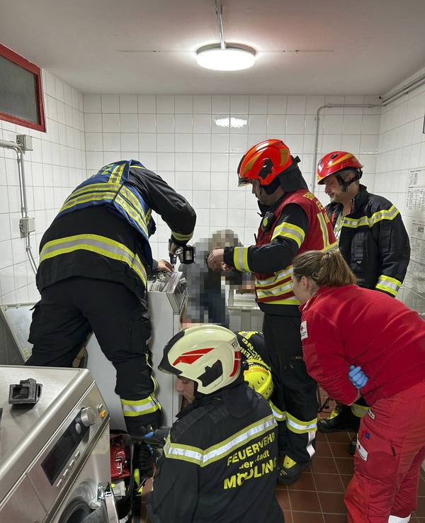 Vier Feuerwehrmänner und eine Rettungssanitäterin sind in einem gekachelten Raum im Einsatz, ein Wehrmann hilft einer Person aus einem Wäschetrockner.