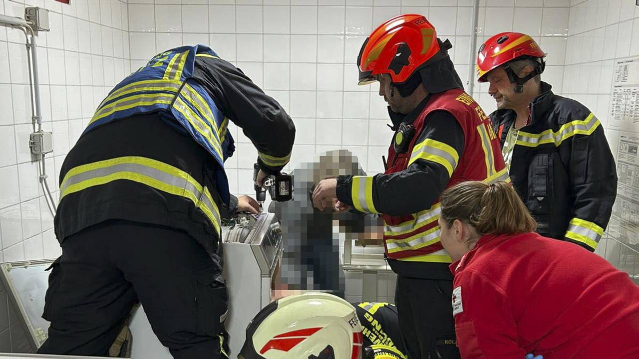 Vier Feuerwehrmänner und eine Rettungssanitäterin sind in einem gekachelten Raum im Einsatz, ein Wehrmann hilft einer Person aus einem Wäschetrockner.
