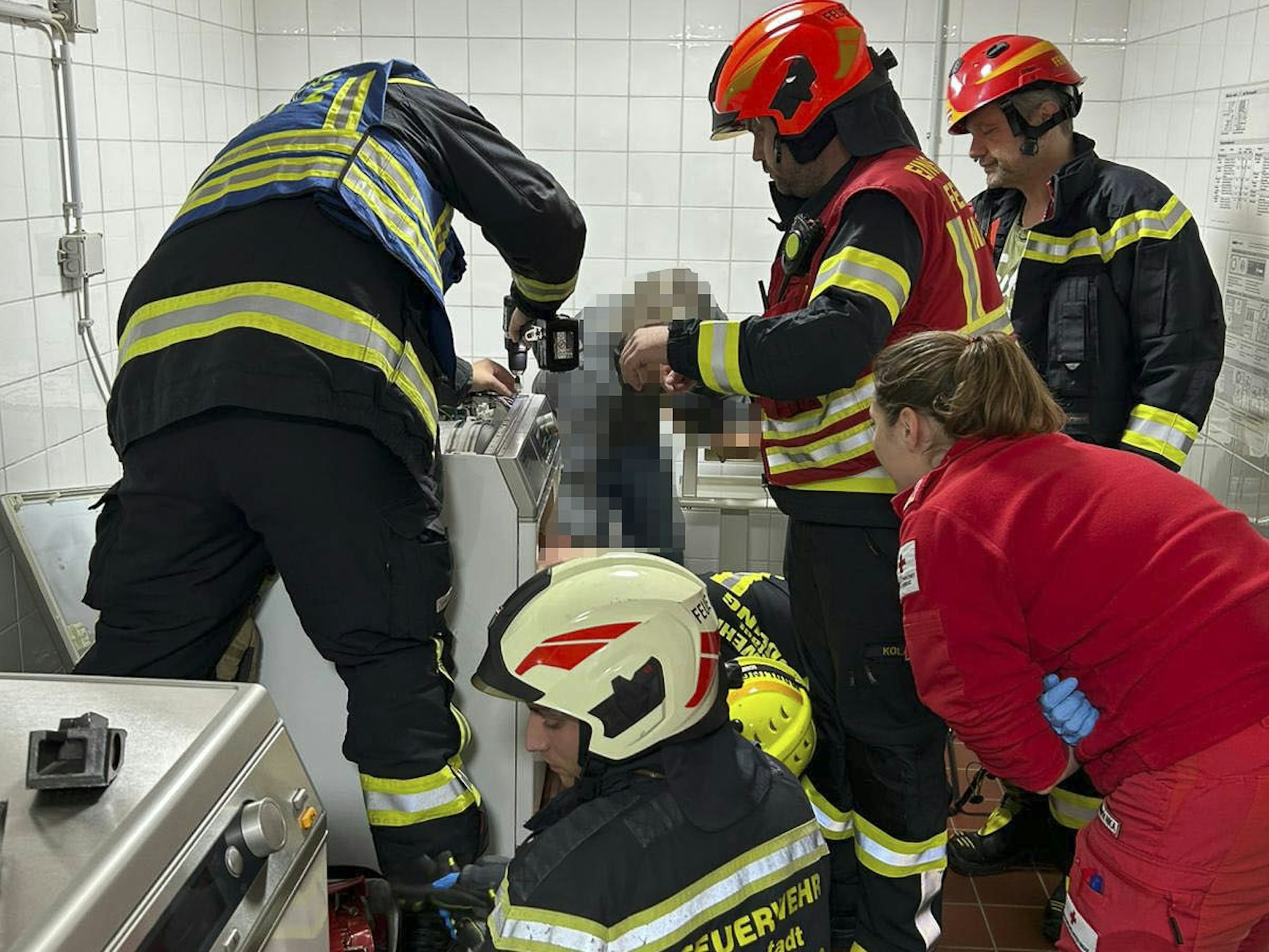 Vier Feuerwehrmänner und eine Rettungssanitäterin sind in einem gekachelten Raum im Einsatz, ein Wehrmann hilft einer Person aus einem Wäschetrockner.