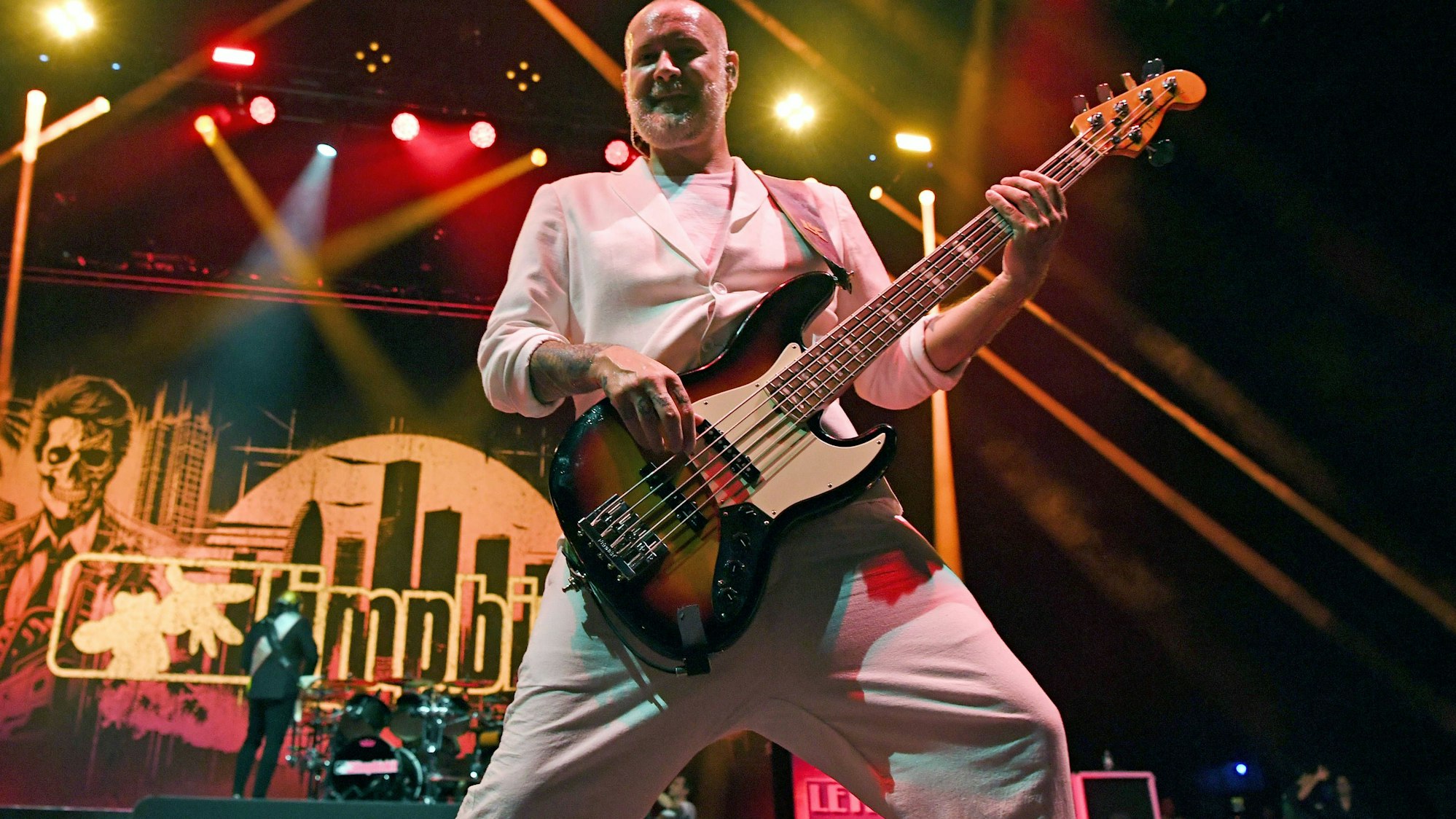 Sam Rivers als Bassist in weißem Anzug auf der Bühne mit Lichtern und Totenkopf-Hintergrund