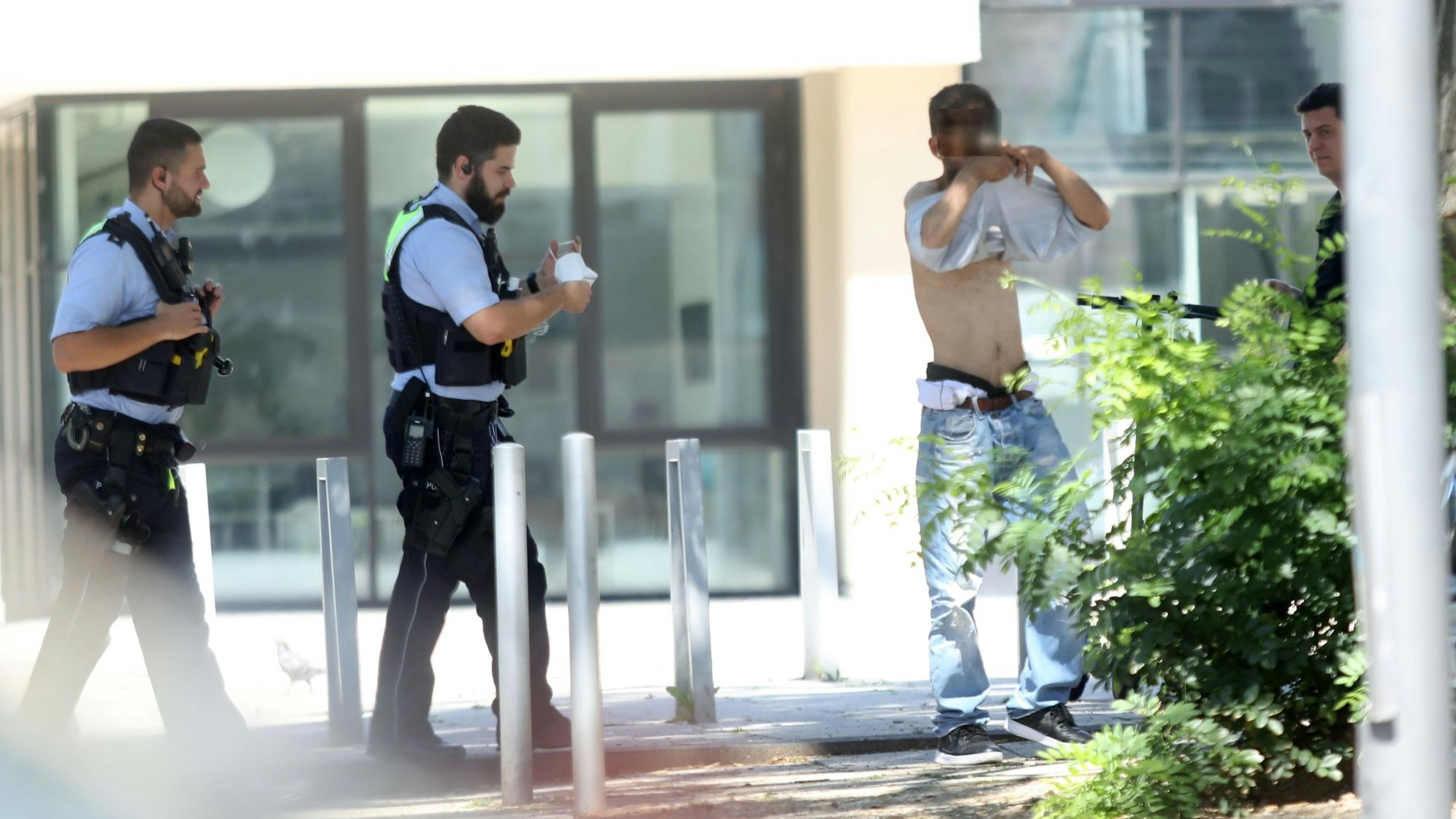 Zwei Polizisten gehen auf einen Mann zu, der vor einem dritten Polizisten sein T-Shirt auszieht. Davor parkt ein Streifenwagen.