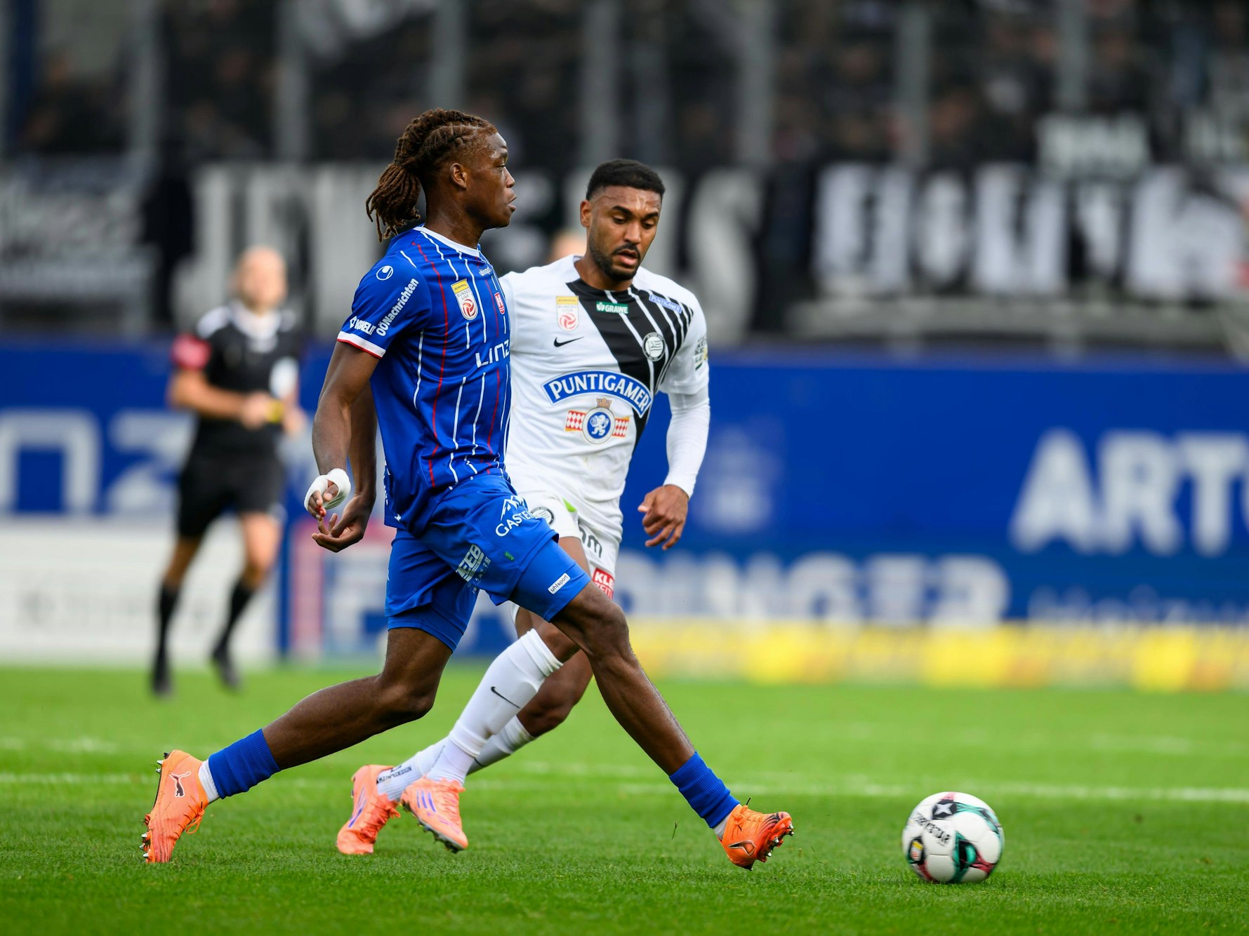 Elias Bakatukanda passt einen Ball, sein Gegenspieler von Sturm Graz steht nicht weit entfernt und schaut auf den Ball.