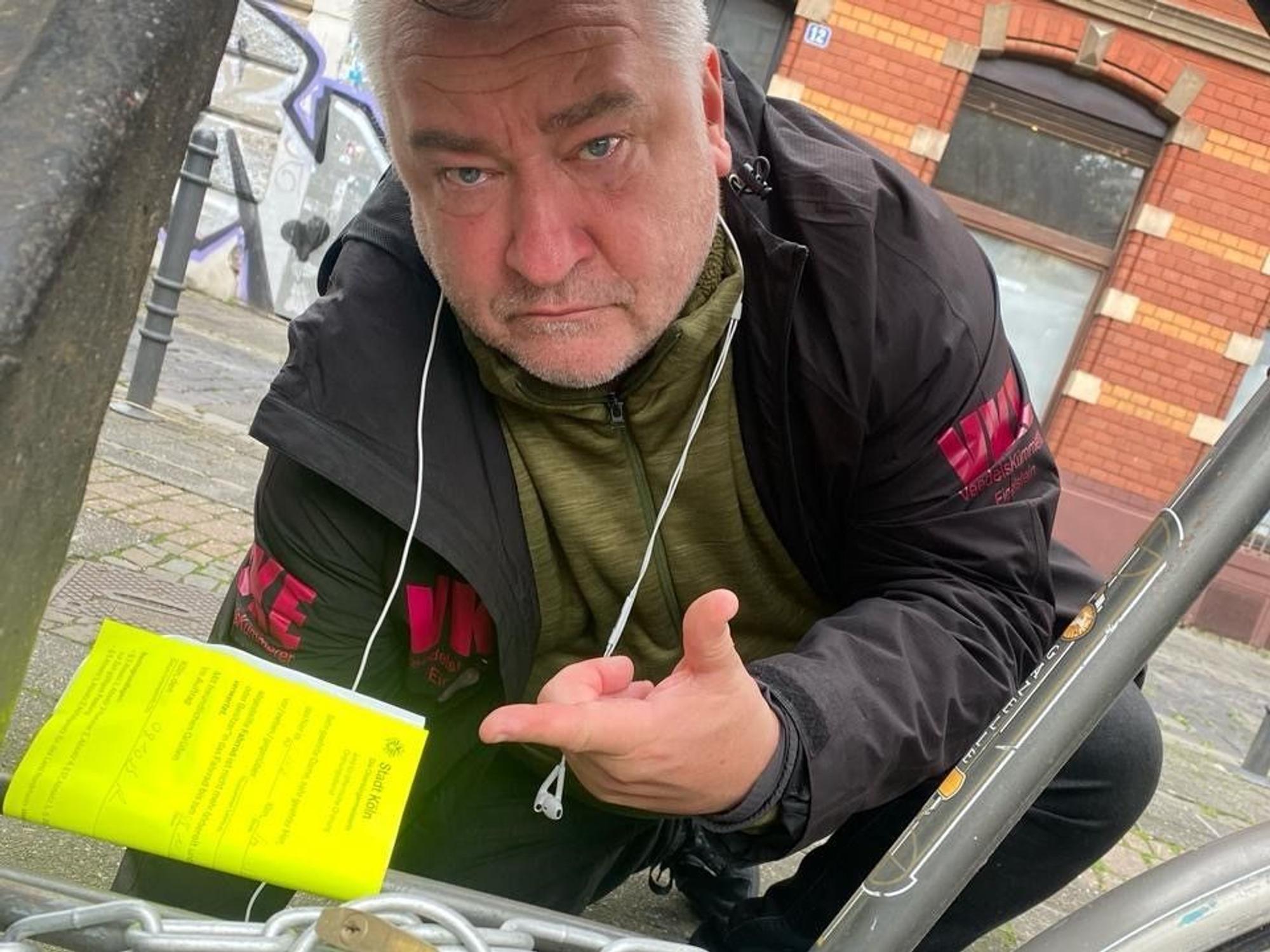 Ein Mann hockt neben einem Fahrrad und zeigt auf einen Zettel, der am Fahrradrahmen befestigt wurde.