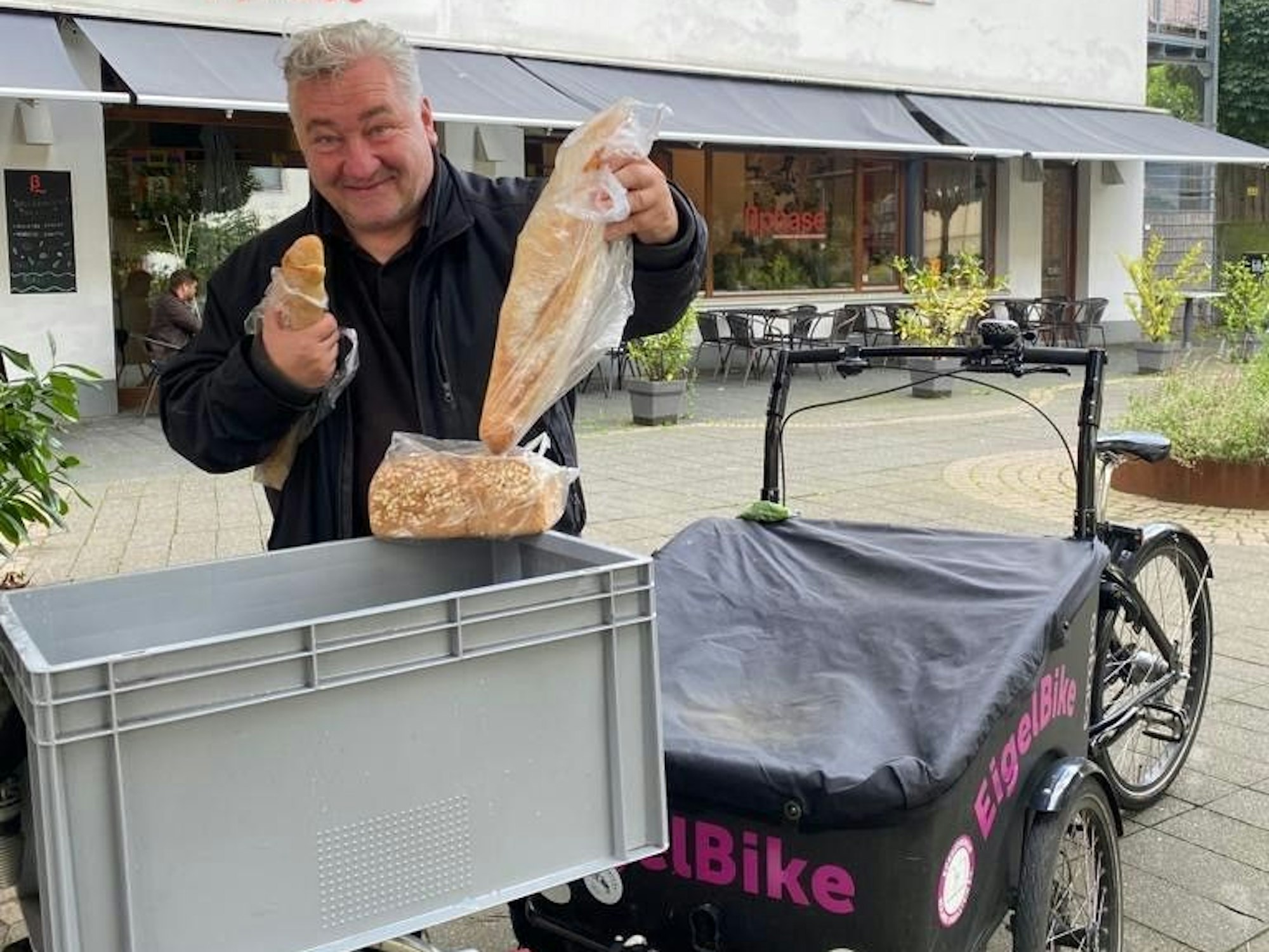 Ein Mann steht an einer Kiste und hält zwei Baguette in der Hand, auf der Kiste liegt ein Brot.