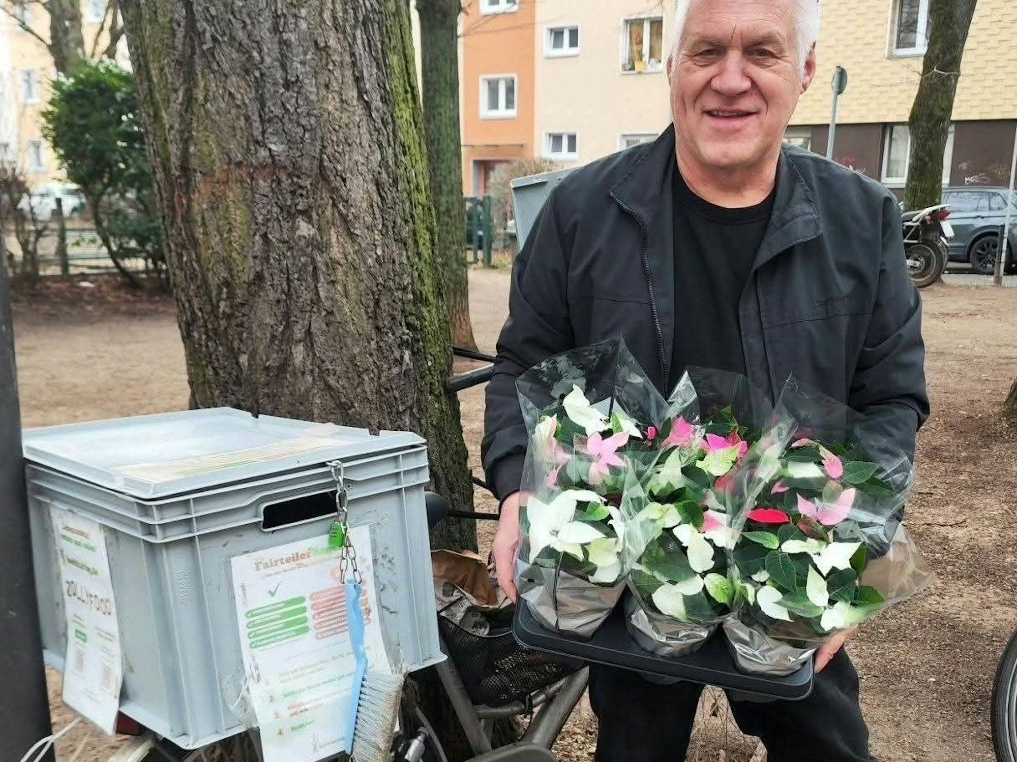 Ein Mann hält mehrere Töpfe mit Blumen in der Hand, er steht neben einem Fahrrad-Fairteiler.