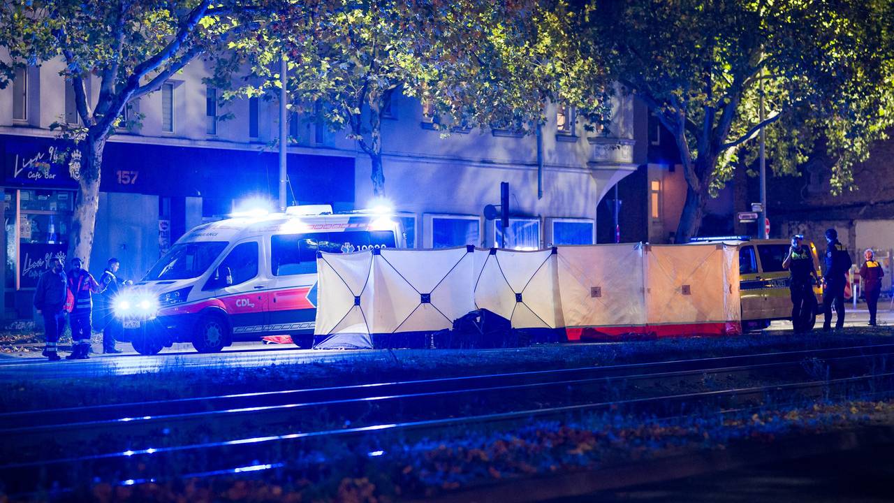 An eine Straße steht ein langer Sichtschutz, dahinter befinden sich mehrere Einsatzkräfte sowie ein Rettungswagen.