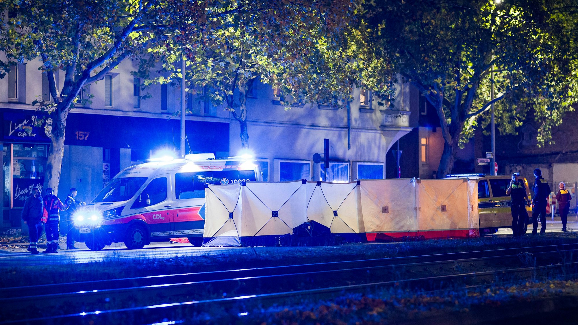 An eine Straße steht ein langer Sichtschutz, dahinter befinden sich mehrere Einsatzkräfte sowie ein Rettungswagen.