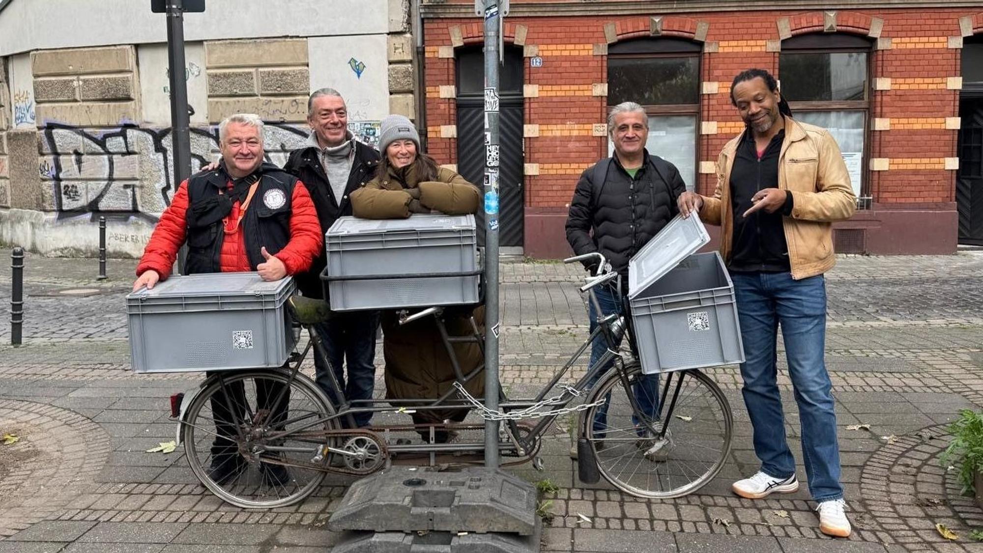 Hinter einem Fahrrad, auf dem sich drei große Plastikkisten befinden, stehen vier Männer und eine Frau.