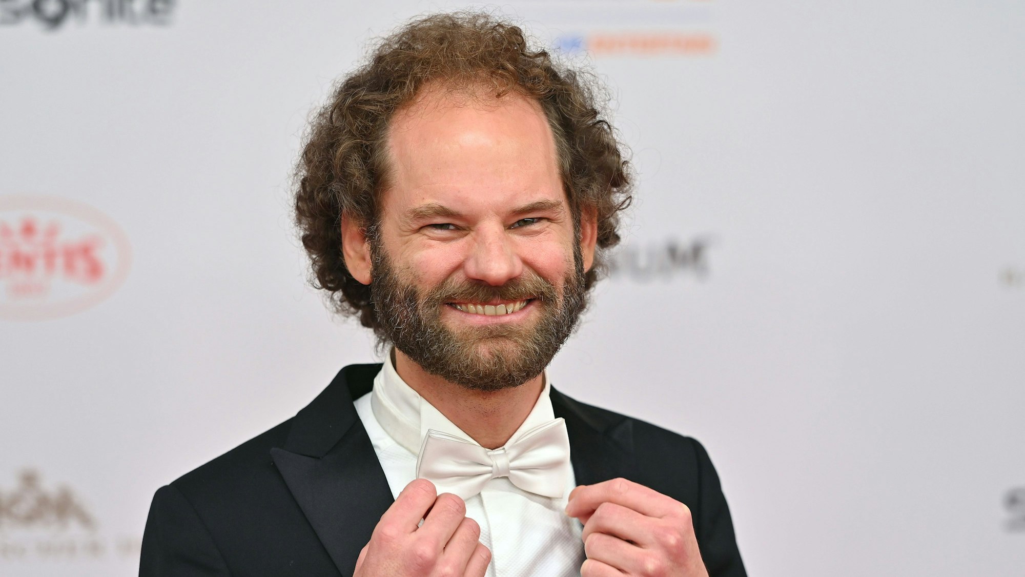 Ein Mann mit Lockenfrisur zupft sich seine weiße Fliege zurecht und lacht.