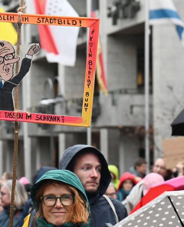 Bei einer Demo hält eine Frau einen Papp-Bilderrahmen hoch, in dem ein Papp-Mann mit dem Gesicht von Bundeskanzler Friedrich Merz ist.