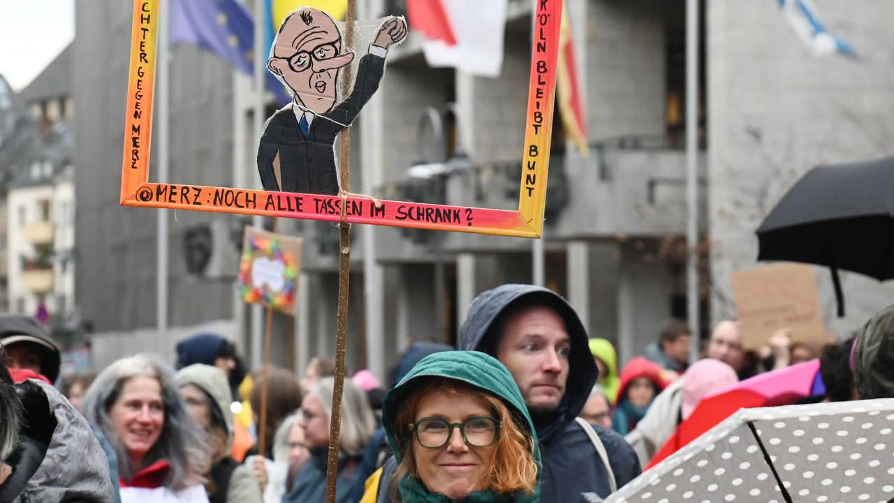 Bei einer Demo hält eine Frau einen Papp-Bilderrahmen hoch, in dem ein Papp-Mann mit dem Gesicht von Bundeskanzler Friedrich Merz ist.