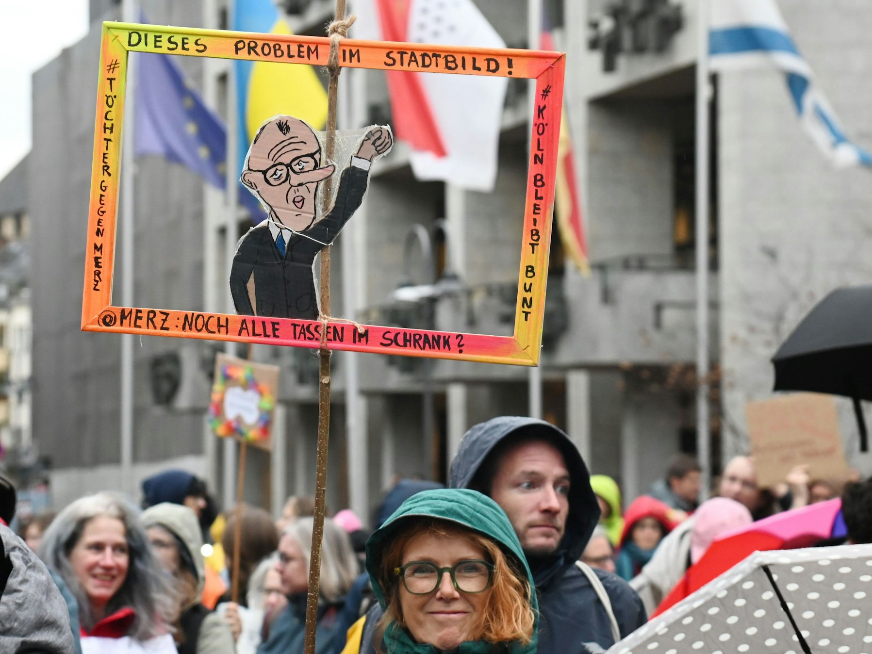 Bei einer Demo hält eine Frau einen Papp-Bilderrahmen hoch, in dem ein Papp-Mann mit dem Gesicht von Bundeskanzler Friedrich Merz ist.