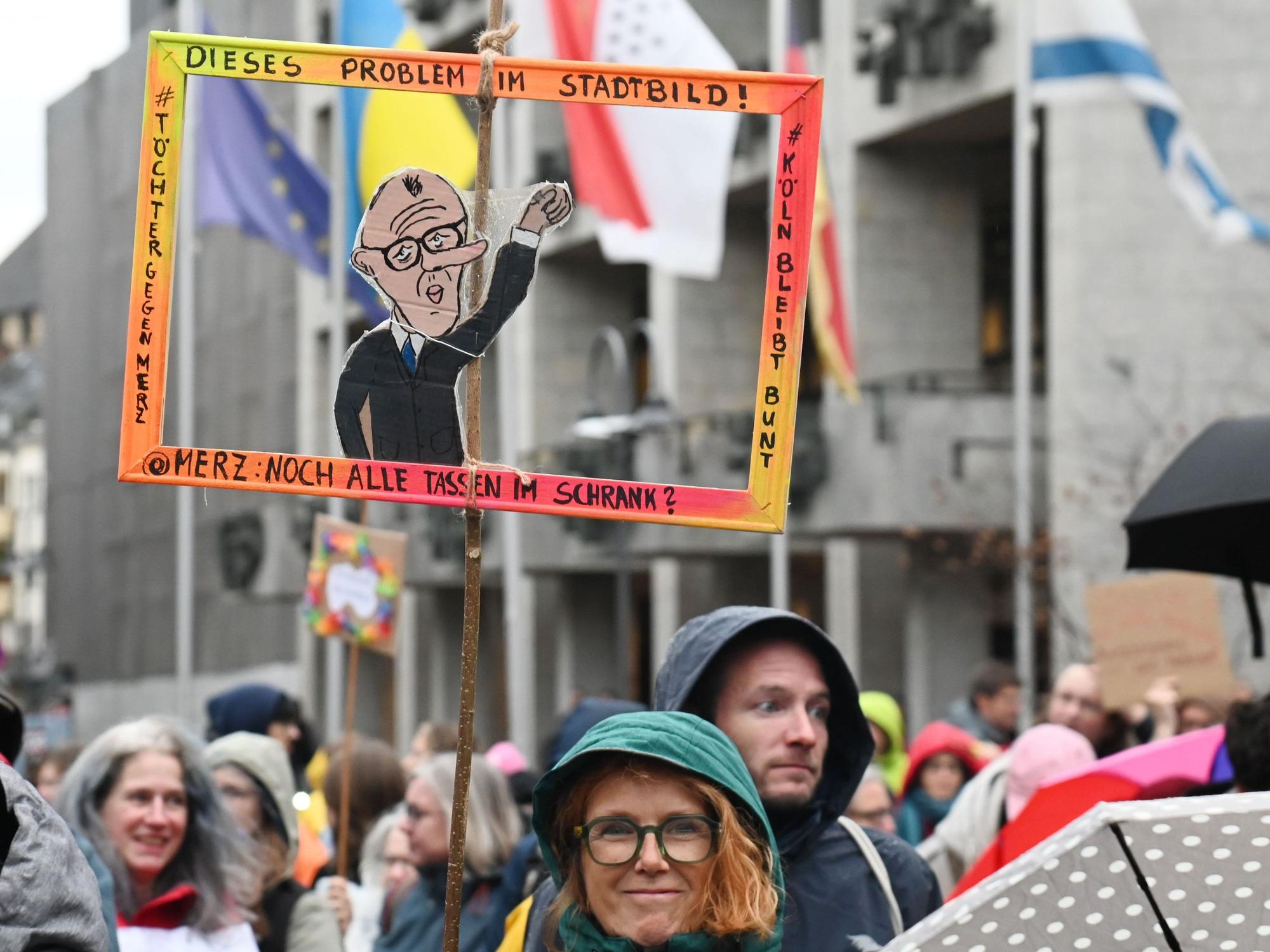 Bei einer Demo hält eine Frau einen Papp-Bilderrahmen hoch, in dem ein Papp-Mann mit dem Gesicht von Bundeskanzler Friedrich Merz ist.