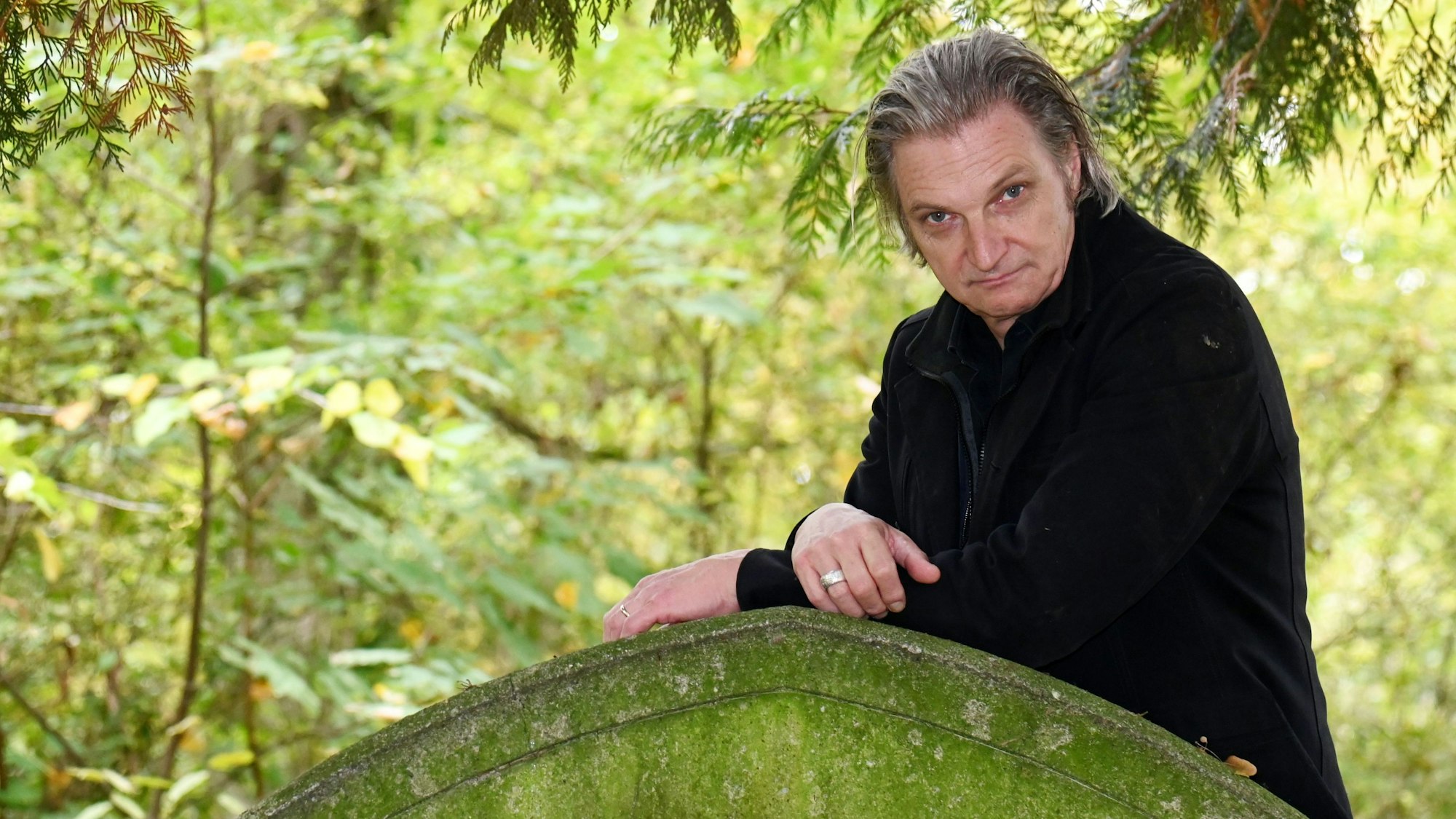 Ein Mann mit mittellangen, graumelierten Haaren lehnt an einem Grabstein.