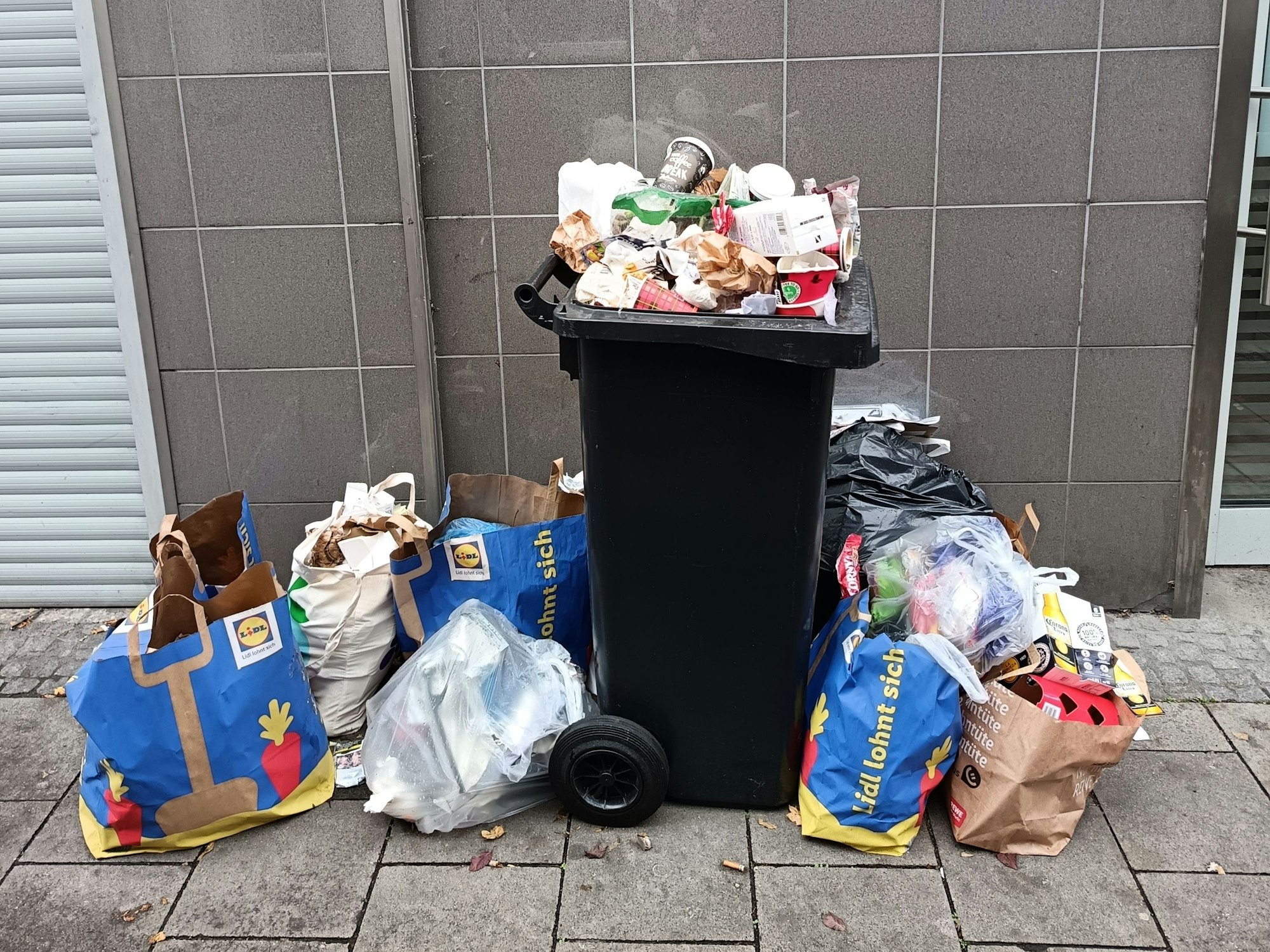 Eine Mülltonne quillt über, um sie herum stehen dazu noch mehrere volle Mülltüten auf dem Boden.