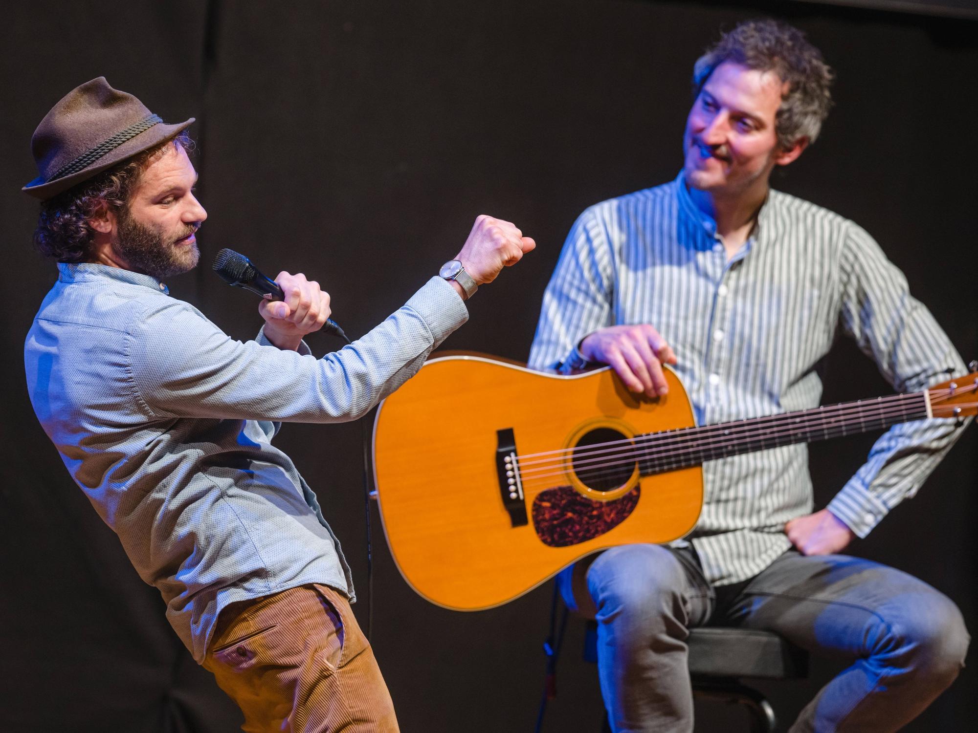 Ein Mann mit Hut hält ein Mikrofon und singt, während ein Mann mit Gitarre auf einem Hocker sitzt