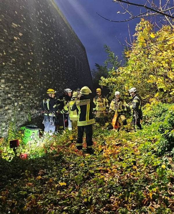 Einsatzkräfte der Feuerwehr stehen unterhalb einer hohen Mauer.