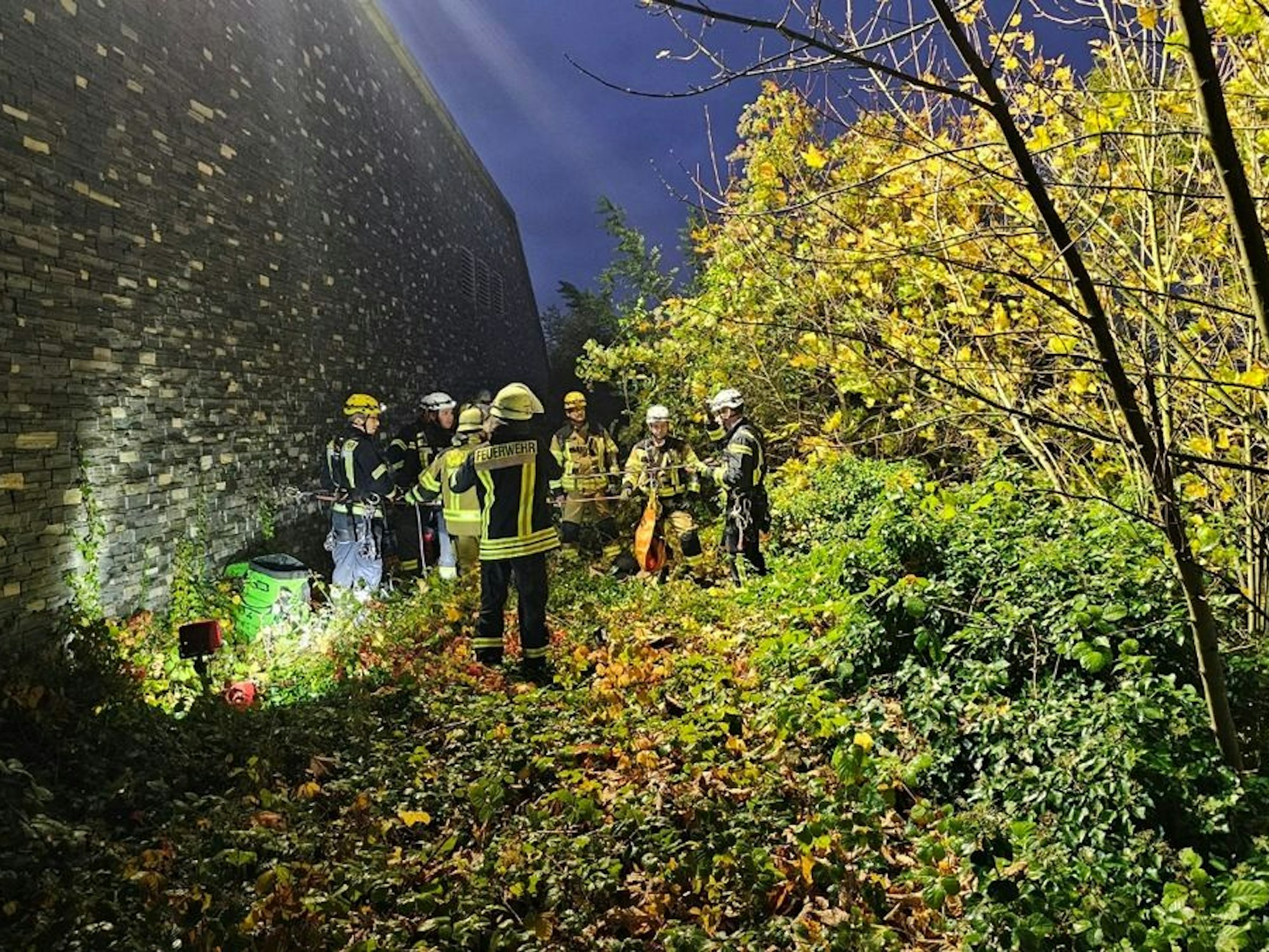 Einsatzkräfte der Feuerwehr stehen unterhalb einer hohen Mauer.