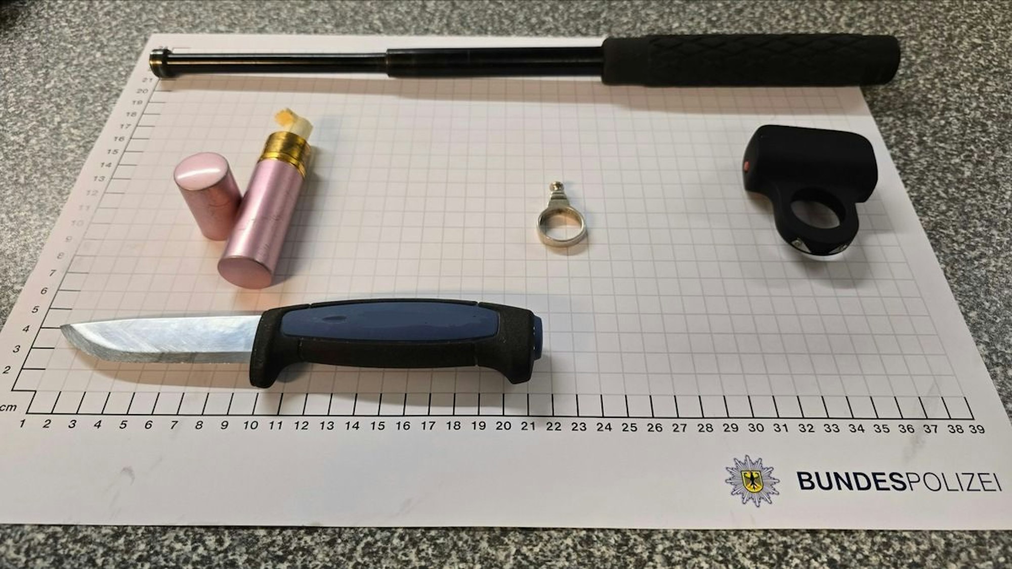 Aufgeklapptes Einhandmesser, Schlagring und als Taschenlampe getarnter Elektroschocker liegen auf einem Tisch.