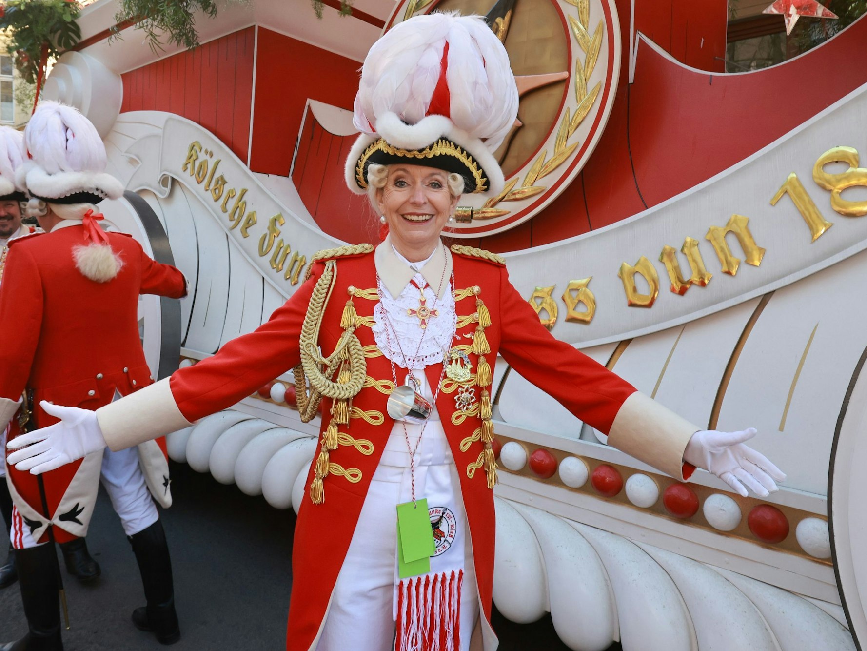 Eine Frau in einer Karnevalsuniform steht vor einem Karnevalswagen und breitet die Arme aus.