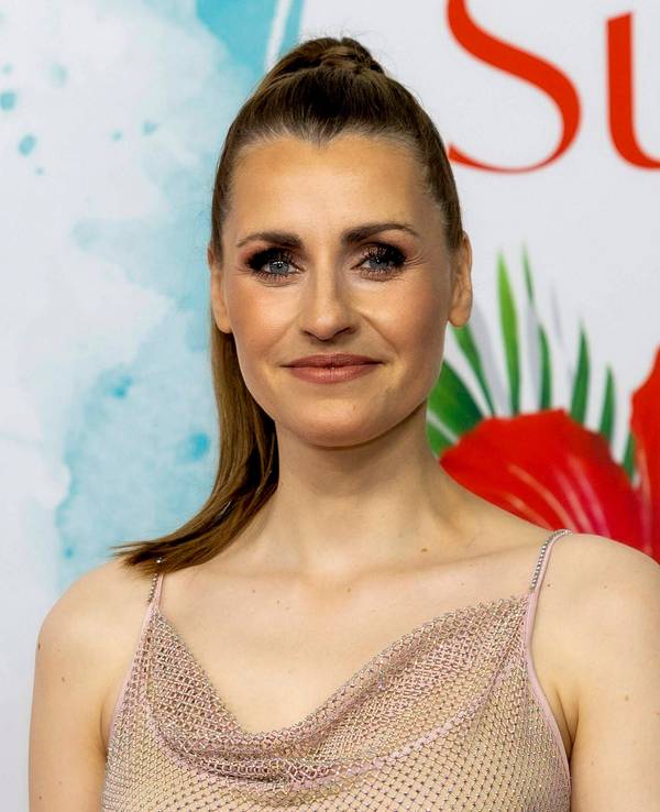 Bella Lesnik mit funkelndem Top lächelt vor einem Hintergrund mit tropischen Blumen und Schrift