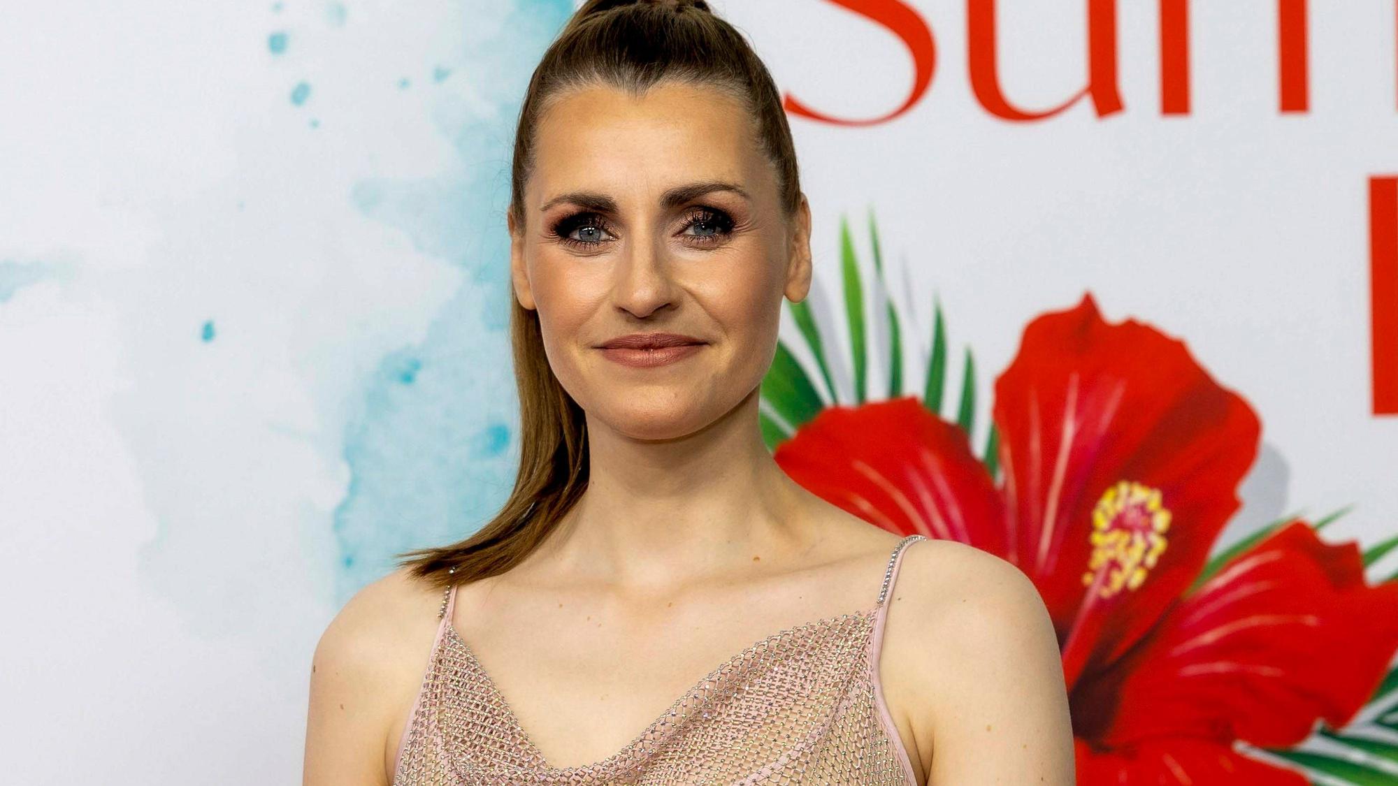 Bella Lesnik mit funkelndem Top lächelt vor einem Hintergrund mit tropischen Blumen und Schrift