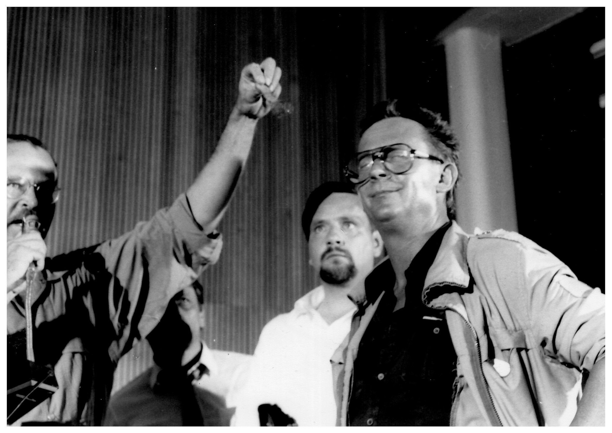 Mehrere Männer stehen auf einer Podiumsbühne, die Person links hat der Person rechts einen falschen Schnäuzer von der Oberlippe gerissen.