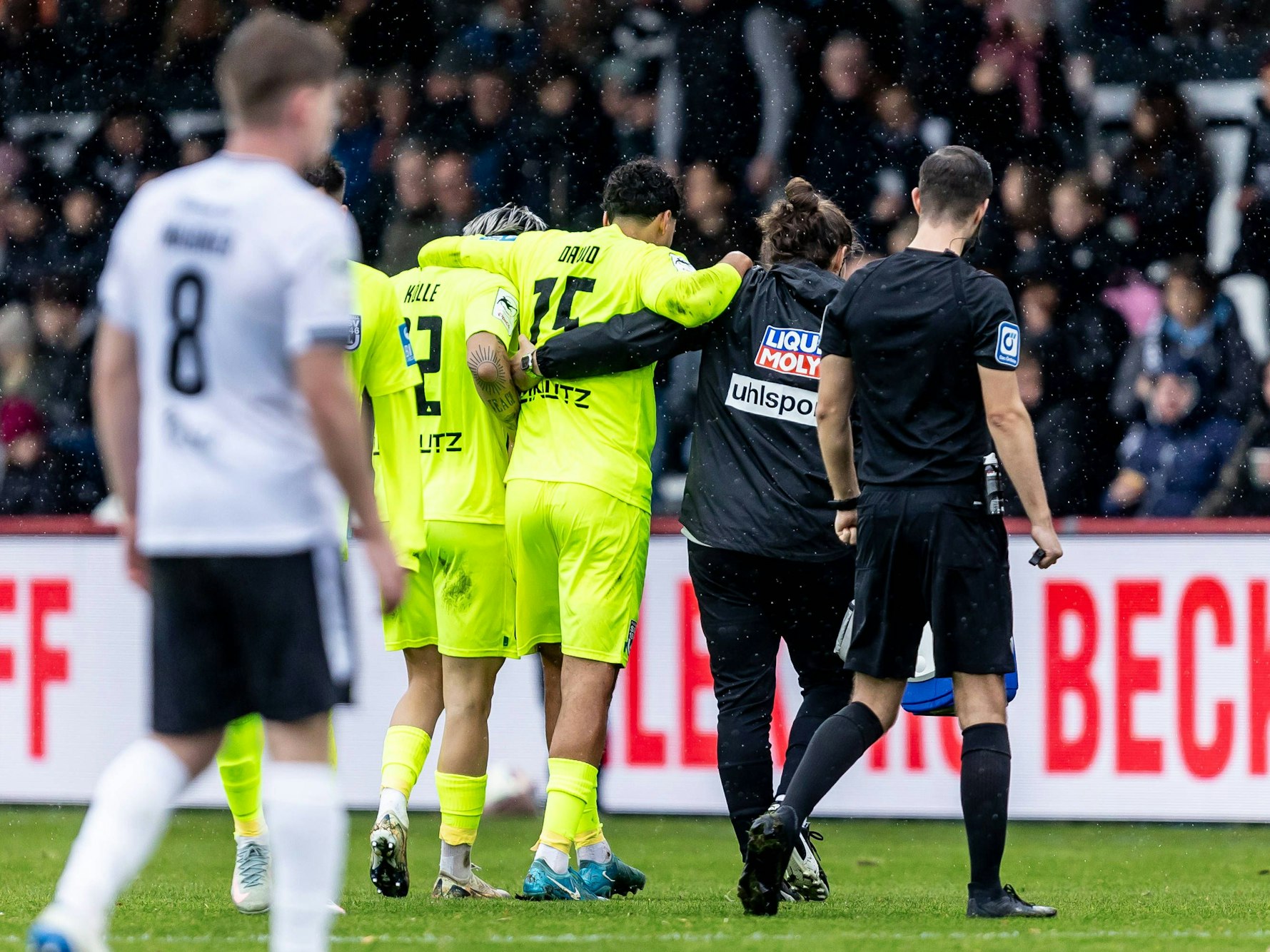 Ein Arzt und ein Mitspieler stützen den verletzten Jonas David und gehen gemeinsam vom Feld.