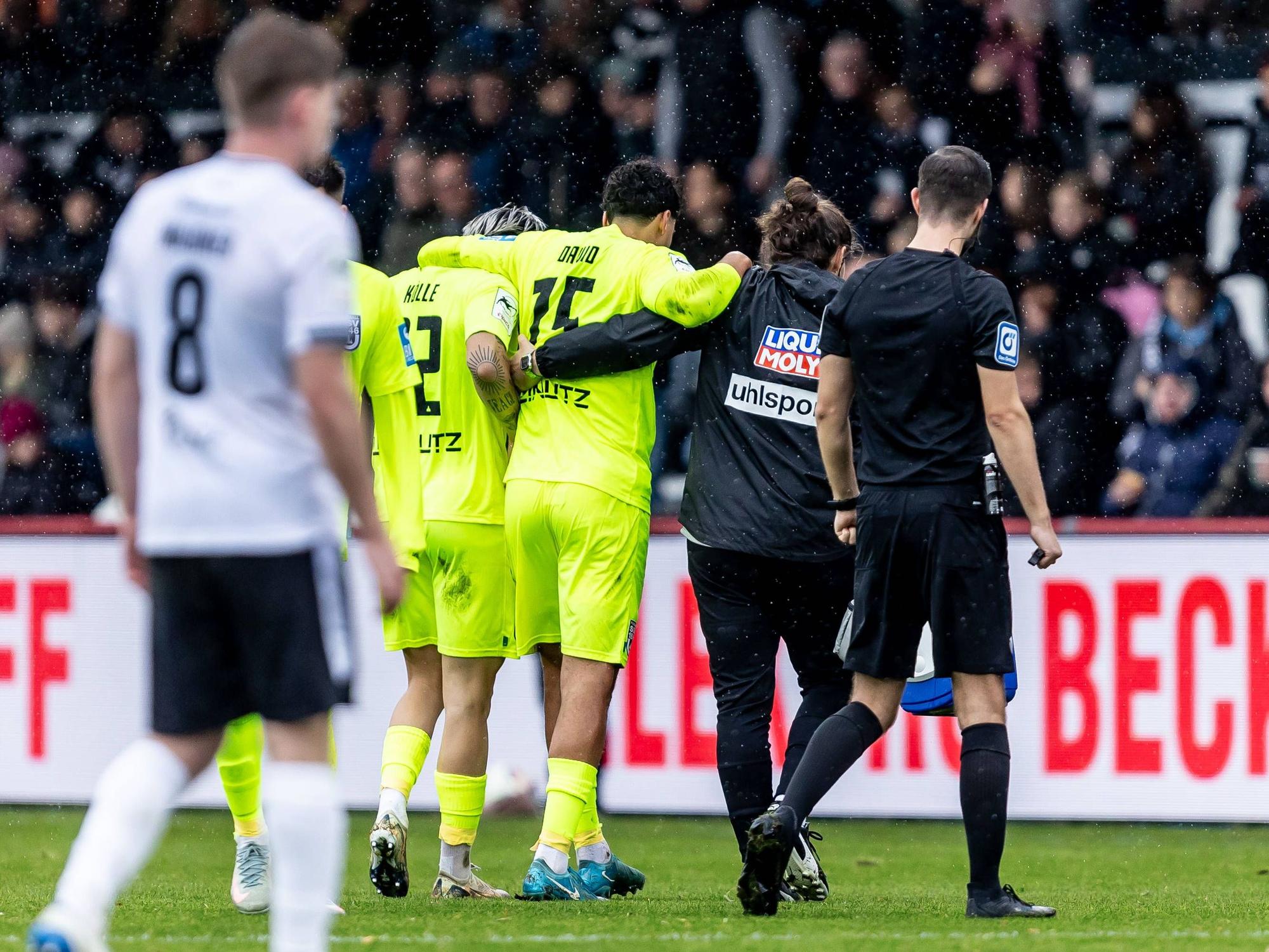 Ein Arzt und ein Mitspieler stützen den verletzten Jonas David und gehen gemeinsam vom Feld.