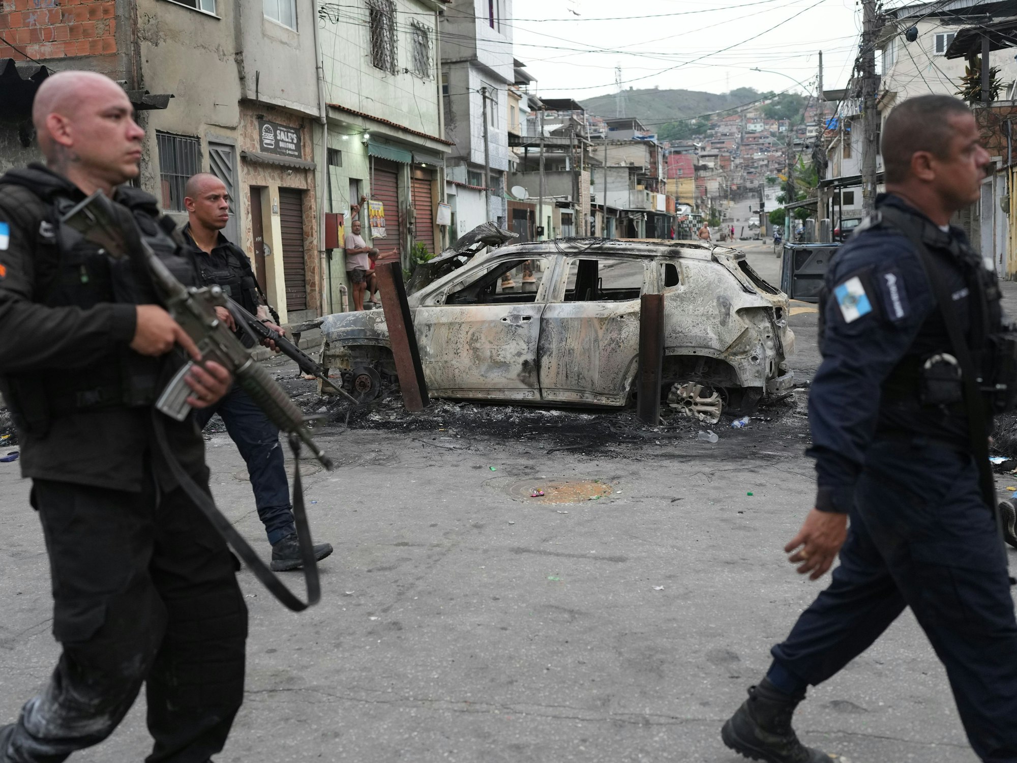 Mit Maschinengewehren bewaffnete Polizisten gehen an einem ausgebrannten Auto vorbei.