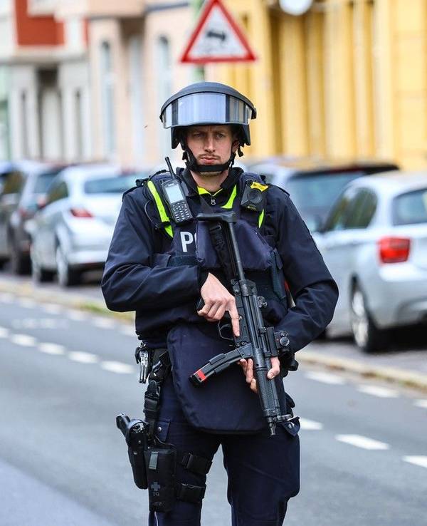 Ein Polizist steht mit einer Maschinenpistole neben einem Streifenwagen an einer Straße.