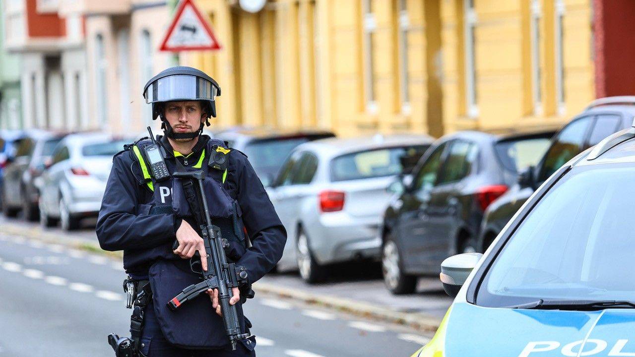 Ein Polizist steht mit einer Maschinenpistole neben einem Streifenwagen an einer Straße.