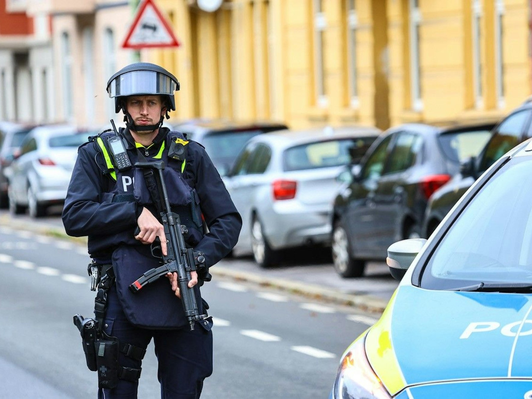 Ein Polizist steht mit einer Maschinenpistole neben einem Streifenwagen an einer Straße.