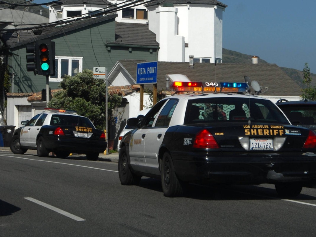 Zwei „Los Angeles County Sheriff“ Autos auf einer Straße mit Häusern im Hintergrund