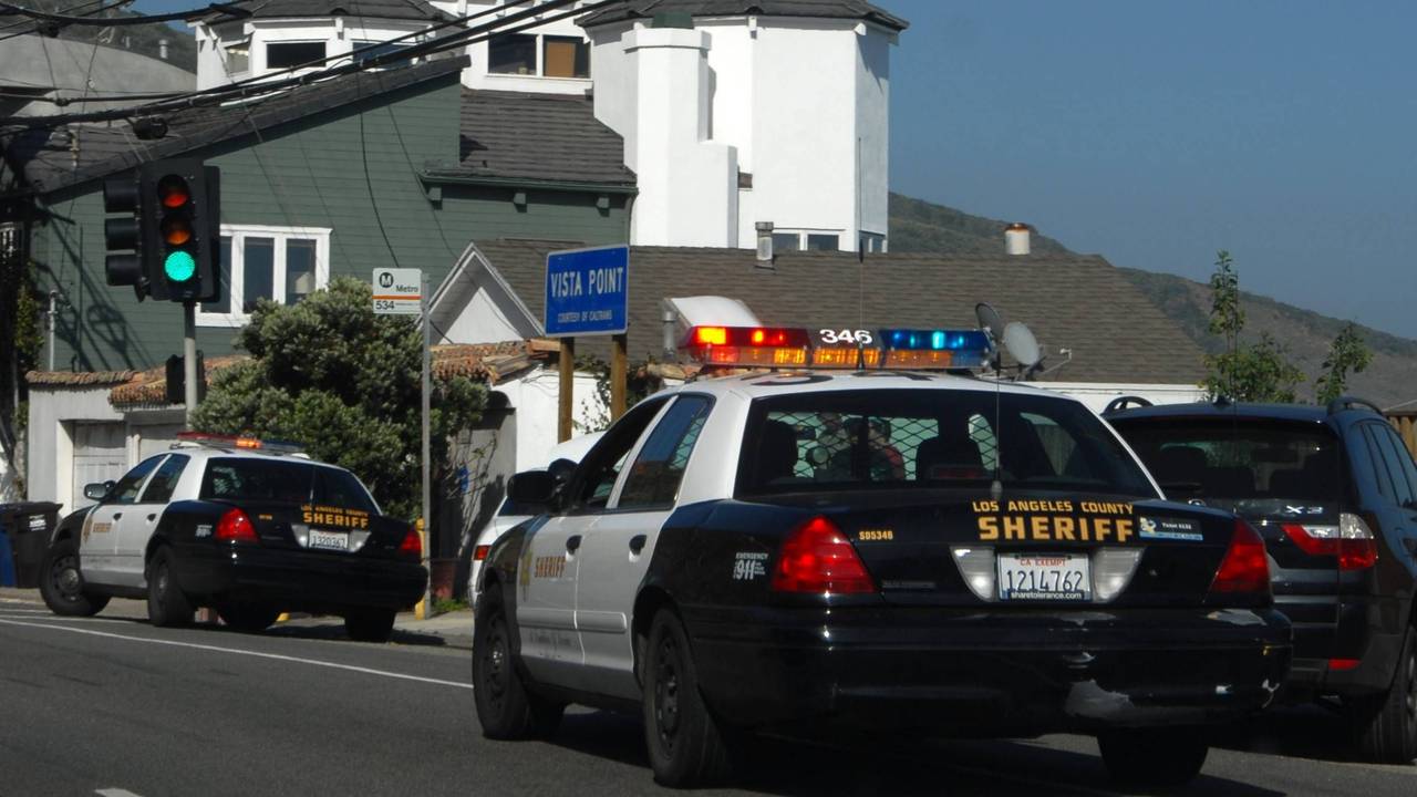 Zwei „Los Angeles County Sheriff“ Autos auf einer Straße mit Häusern im Hintergrund