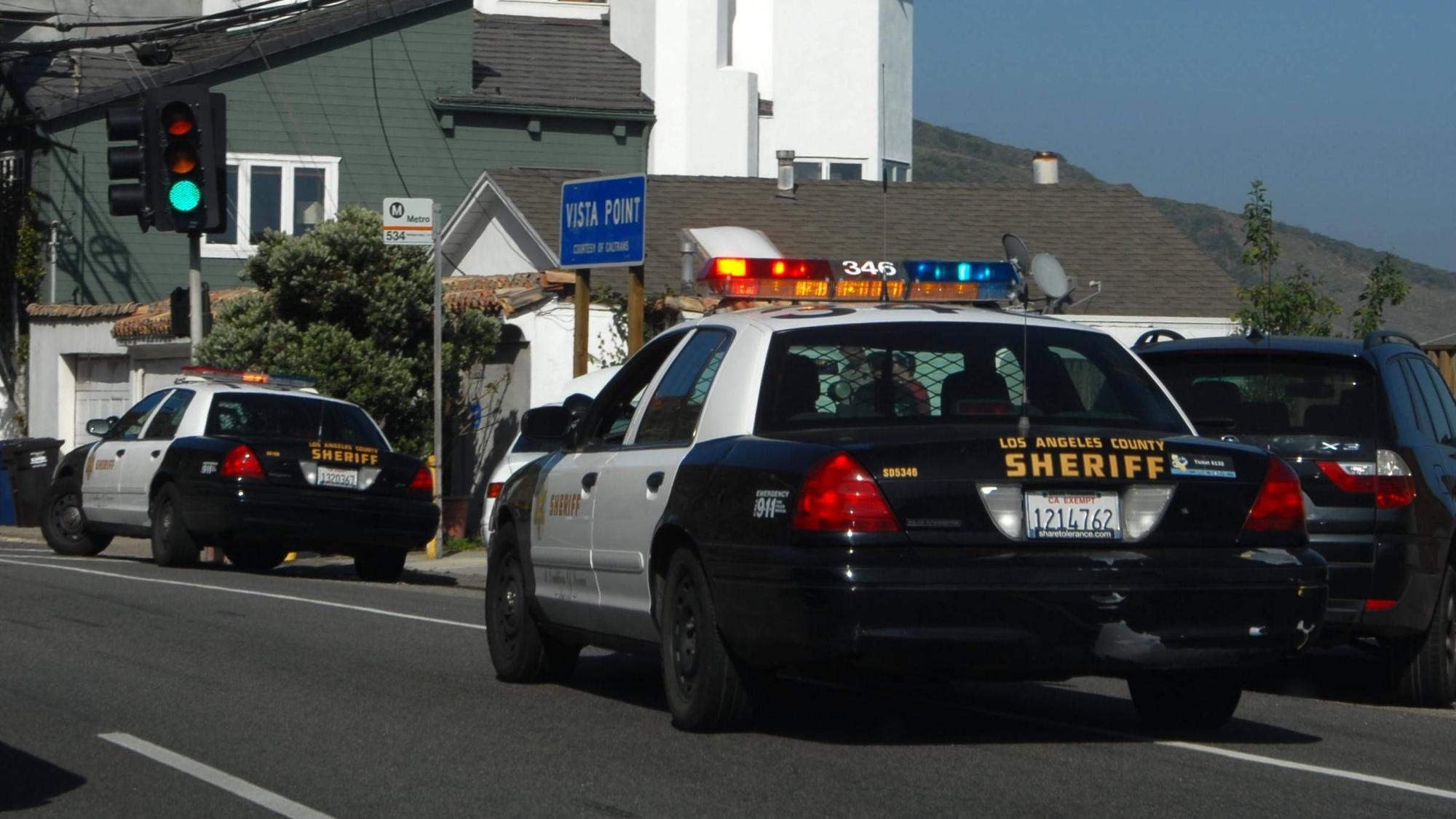 Zwei „Los Angeles County Sheriff“ Autos auf einer Straße mit Häusern im Hintergrund.