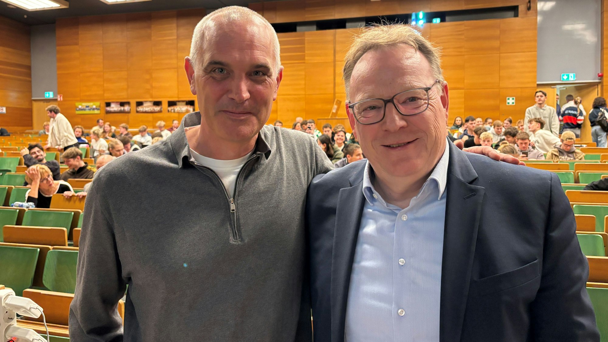 Zwei Männer stehen Arm in Arm in einem Uni-Hörsaal.