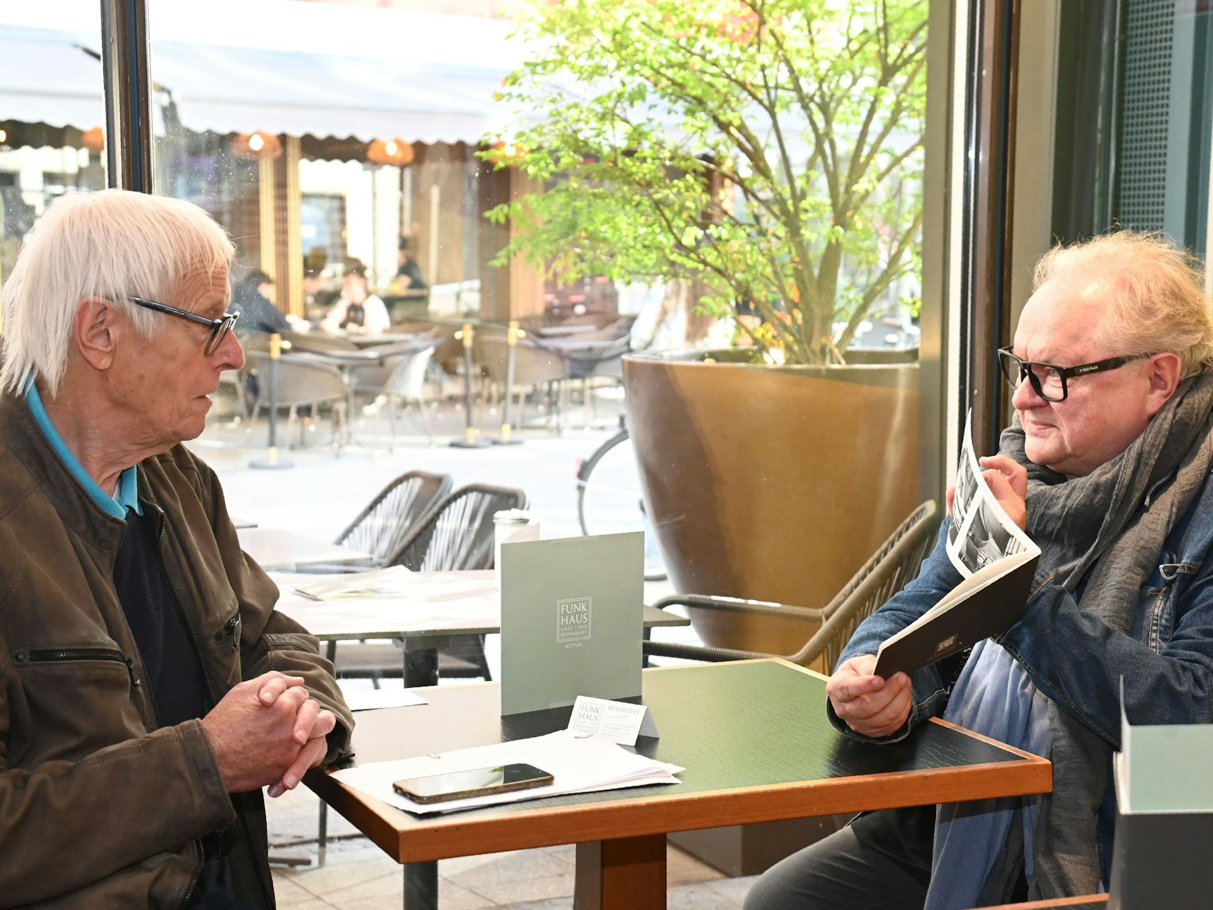 Zwei ältere Herren sitzen an einem Kaffeehaustisch, der eine zeigt dem anderen einen Prospekt.