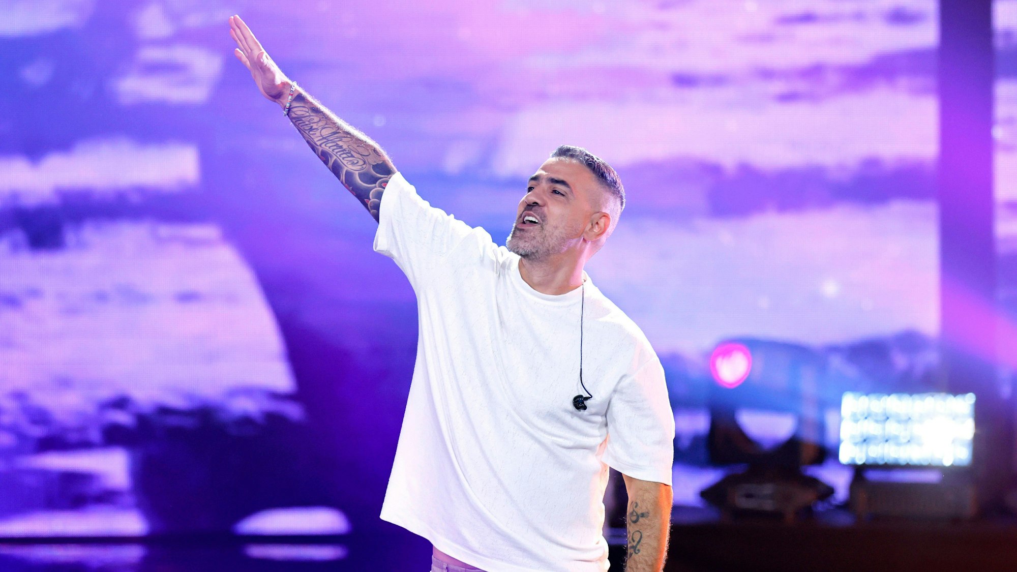 Ein Mann im weißen T-Shirt reckt auf einer Bühne den rechten, volltätowierten Arm in die Luft.