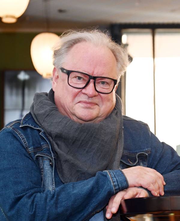 Ein Mann mit Jeansjacke, großem Halstuch und markanter, schwarz umrahmter Brille lehnt an einem Tresen und lächel leicht.