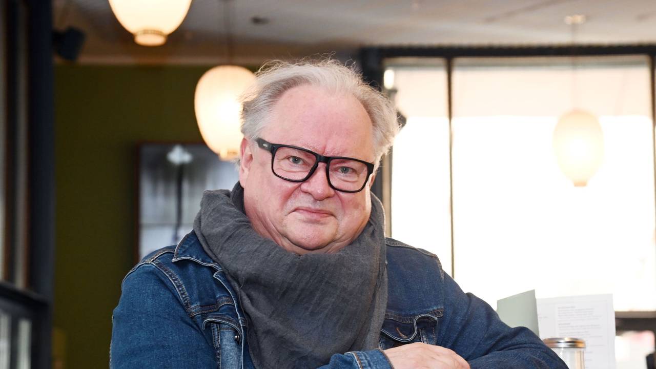 Ein Mann mit Jeansjacke, großem Halstuch und markanter, schwarz umrahmter Brille lehnt an einem Tresen und lächel leicht.