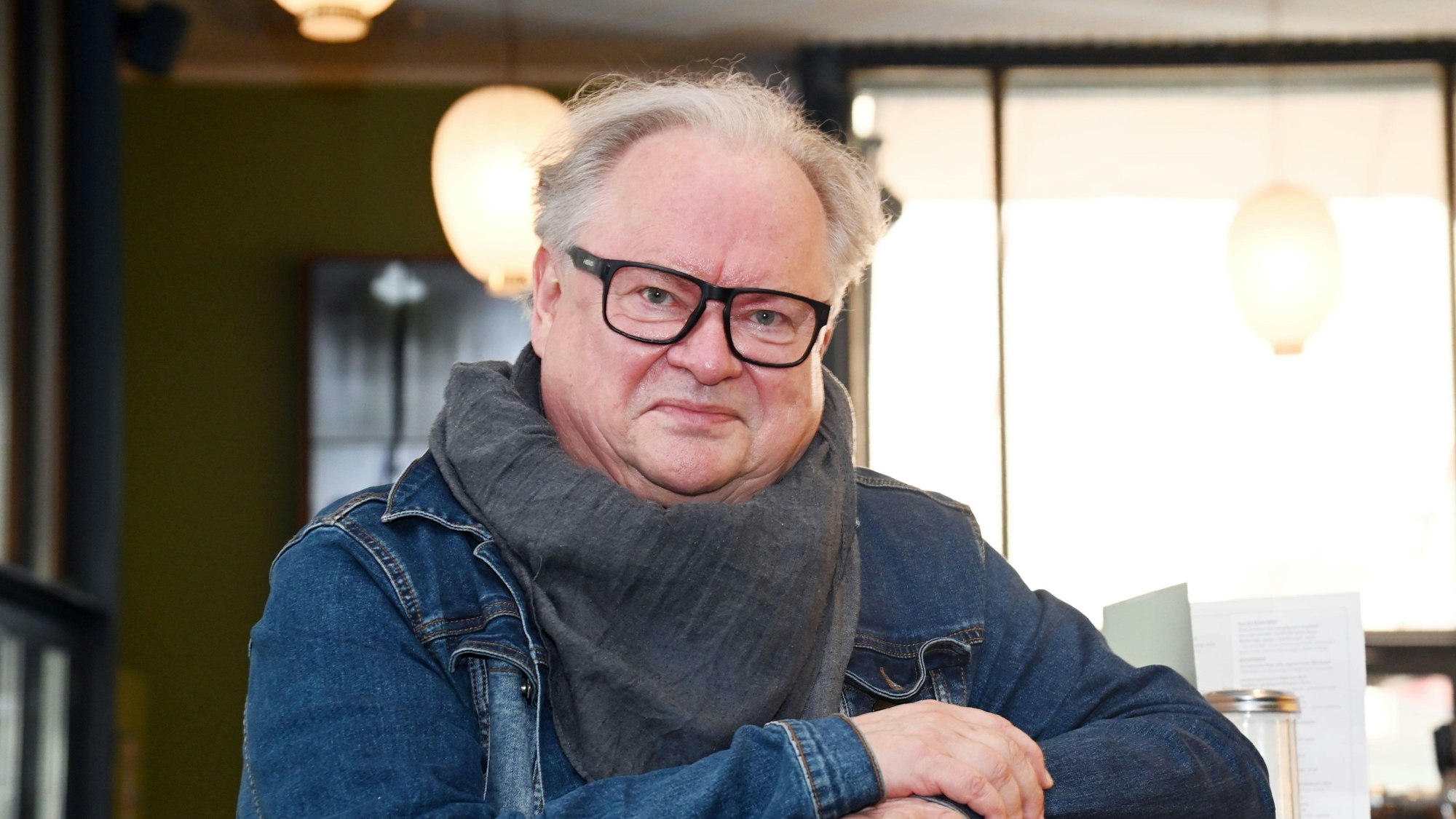 Ein Mann mit Jeansjacke, großem Halstuch und markanter, schwarz umrahmter Brille lehnt an einem Tresen und lächel leicht.