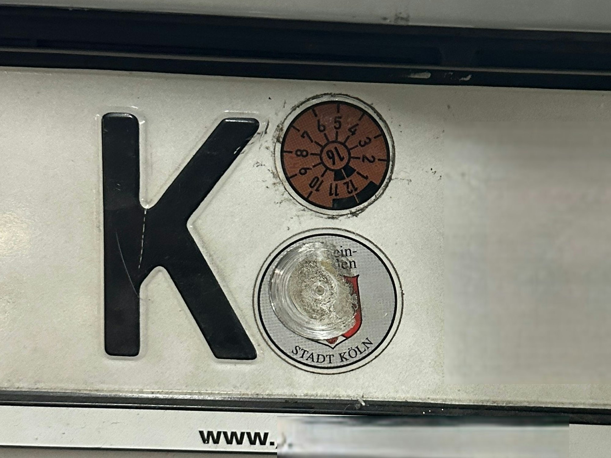 An einem Nummernschild mit Kölner Kennzeichen klebt eine TÜV-Plakette mit einer 16 in der Mitte.